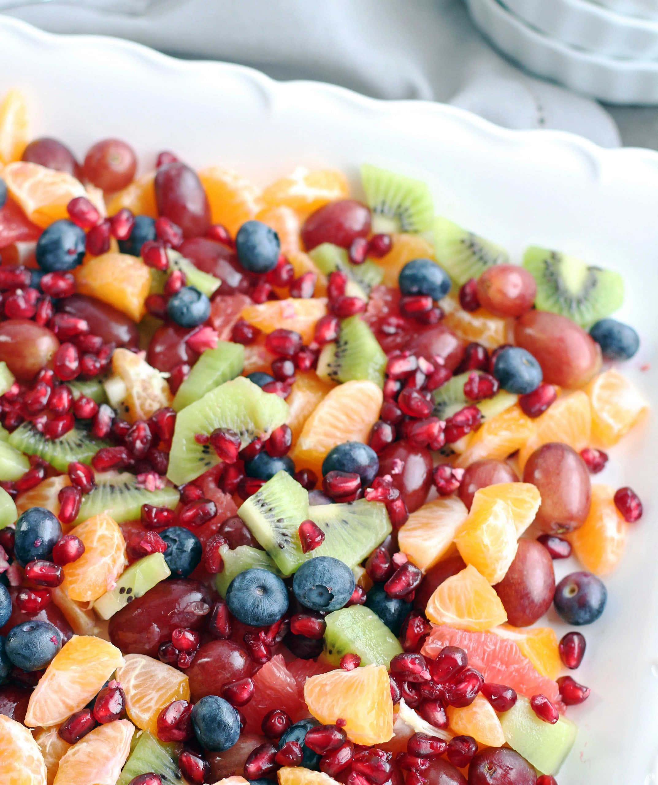 Winter Fruits 桌布 壁紙 — 一個充滿活力的水果沙拉，有石榴籽、奇異果、藍莓和橙瓣，放在一個白色扇貝邊的盤子上。