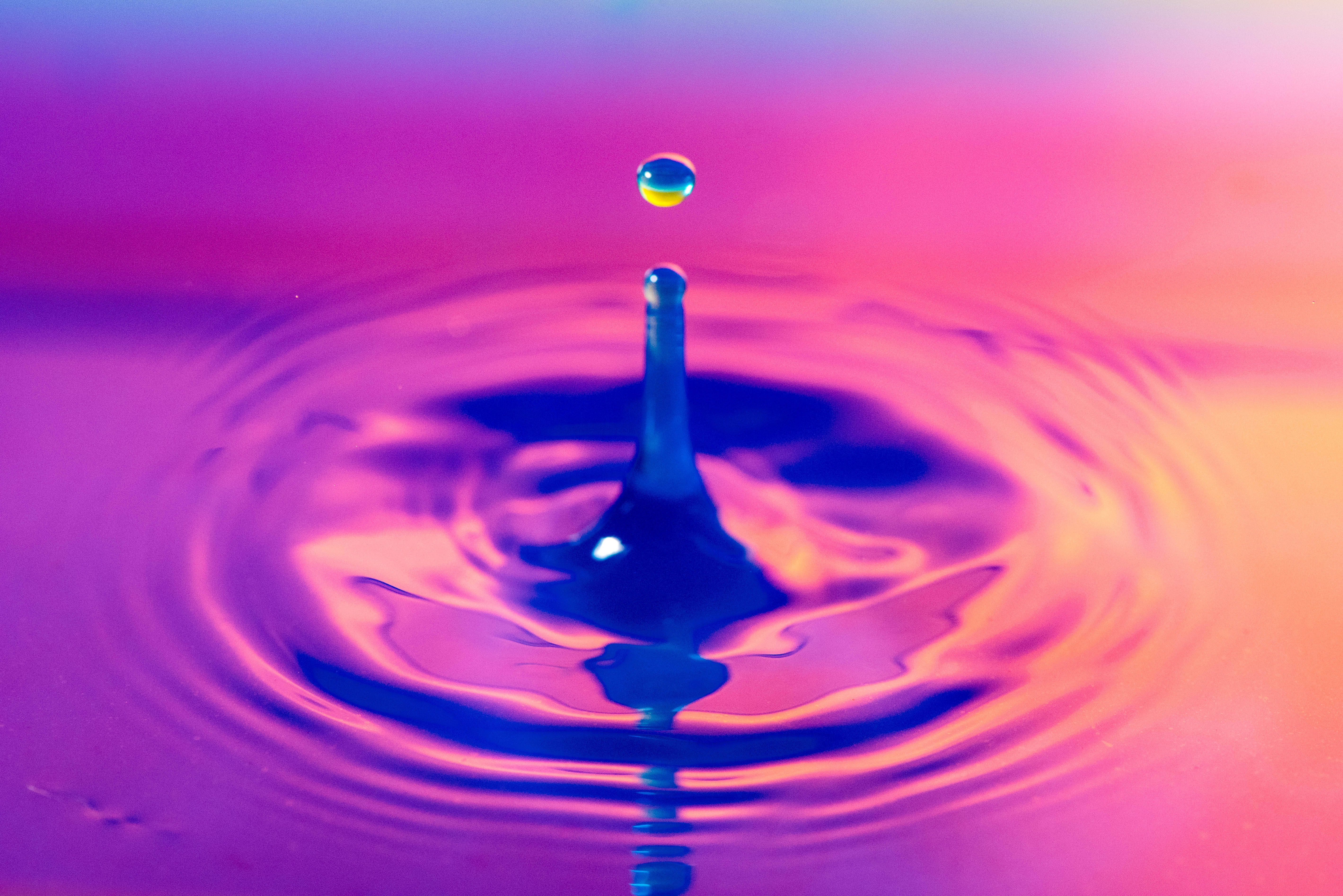 水 桌布 壁紙 — 一個充滿活力的水滴懸浮在一個泛起漣漪的池面上，池中帶有螢光粉紅和藍色調，形成同心圓。