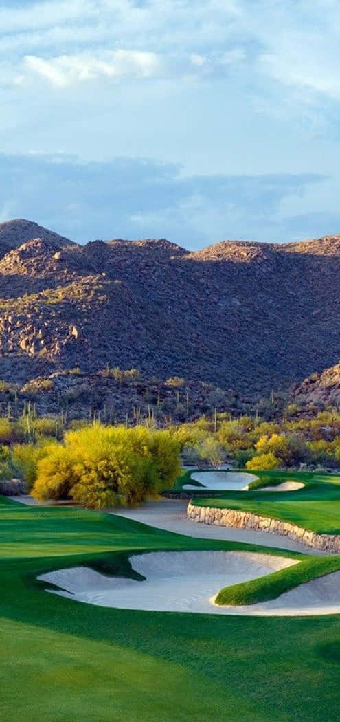 Pixel 3 Golf Course 桌布 壁紙 — 一個充滿活力的沙漠高爾夫球場，擁有鬱鬱蔥蔥的綠色球道和沙坑，坐落在崎嶇的山脈之間，天空部分多雲。