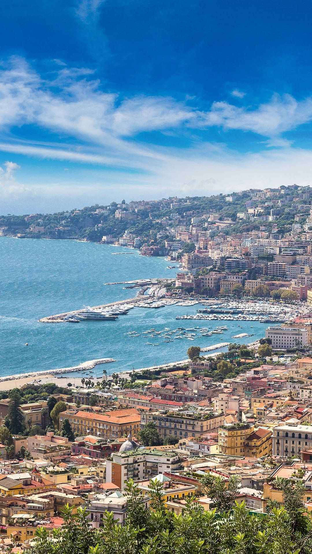 Naples 桌布 壁紙 — 一個充滿活力的沿海城市依附在陽光明媚的山坡上，俯瞰著一個停滿船隻的碧綠海灣，上方是明亮的藍天。