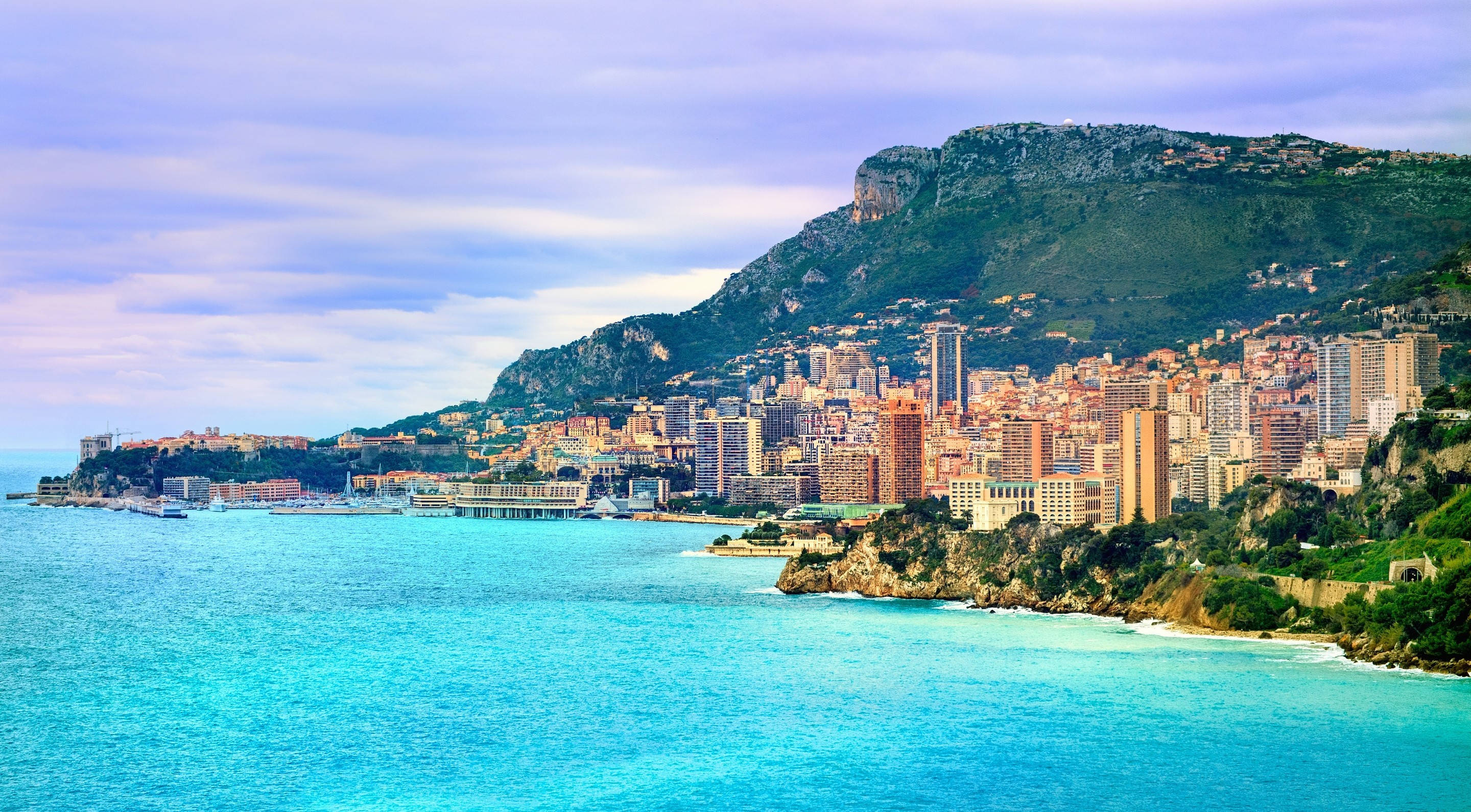 Monaco 桌布 壁紙 — 一個充滿活力的沿海城市依附在鬱鬱蔥蔥的綠色山坡上，俯瞰著柔和的藍天下碧綠的海水。