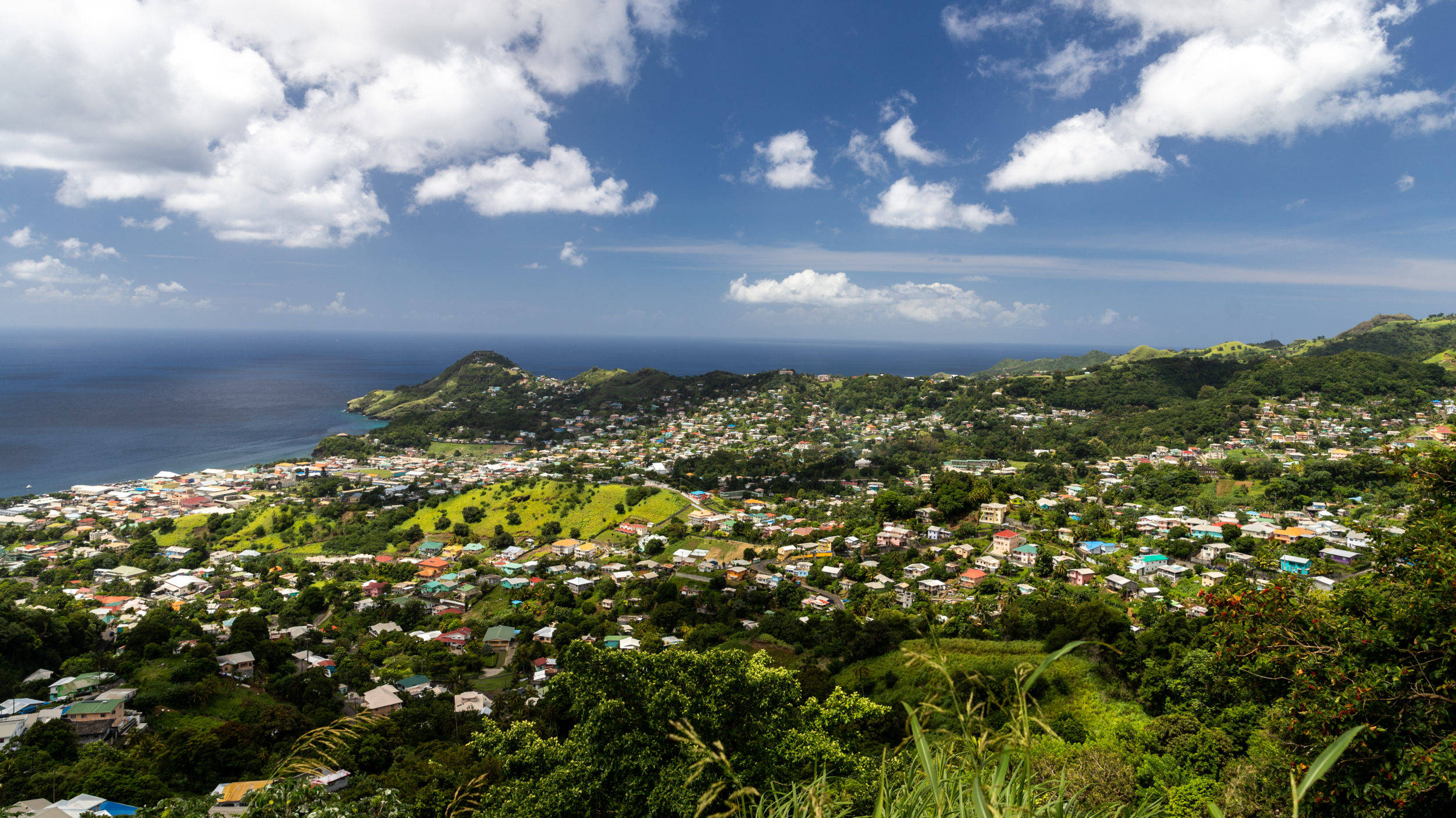 St Vincent And The Grenadines 桌布 壁紙 — 一個充滿活力的沿海小鎮，在點綴著蓬鬆白雲的明亮藍天下，蔓延在鬱鬱蔥蔥的綠色山丘上。
