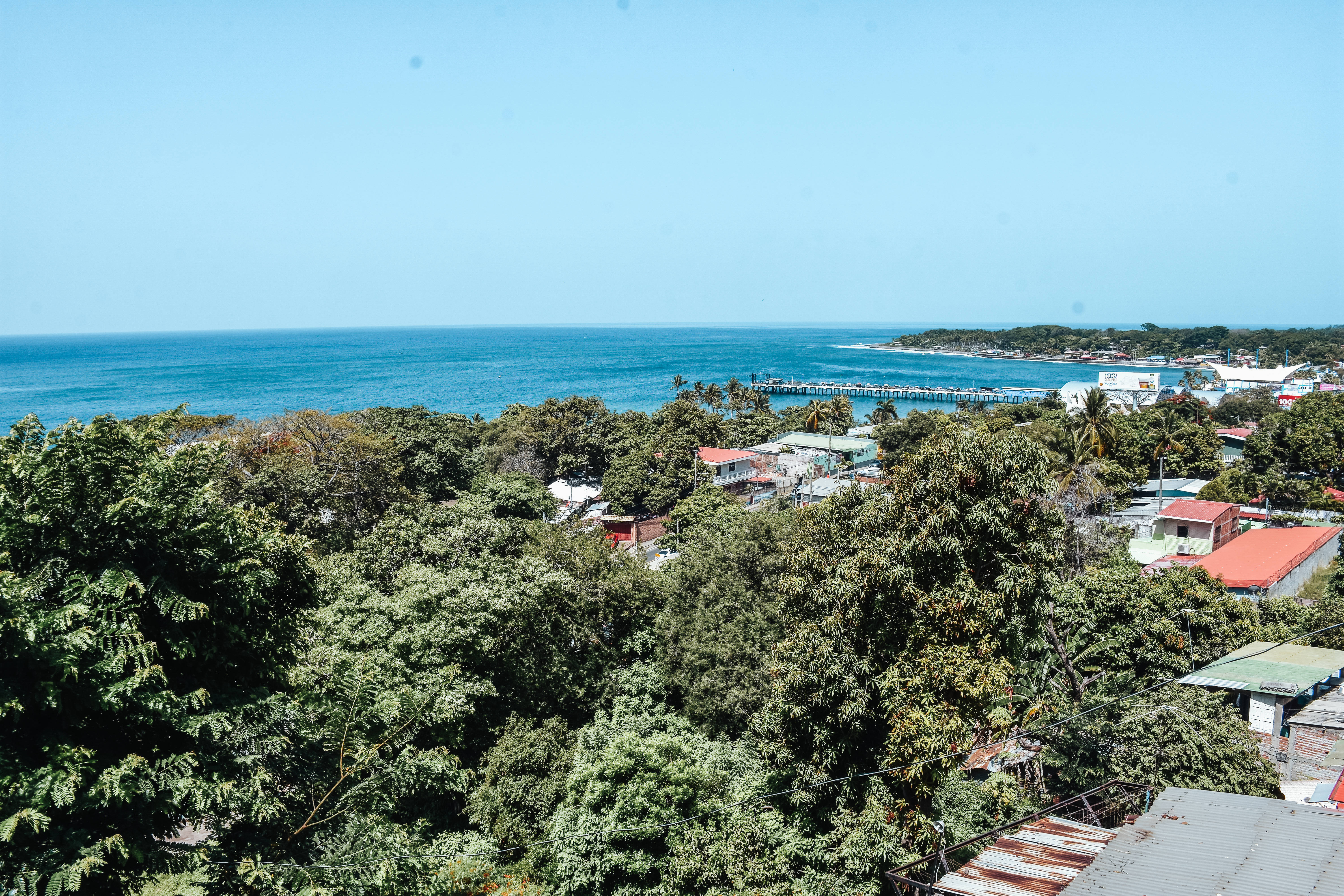 El Salvador 桌布 壁紙 — 一個充滿活力的沿海小鎮，坐落在茂密的綠樹叢中，長長的碼頭在清澈的藍天下延伸至碧綠的海洋。