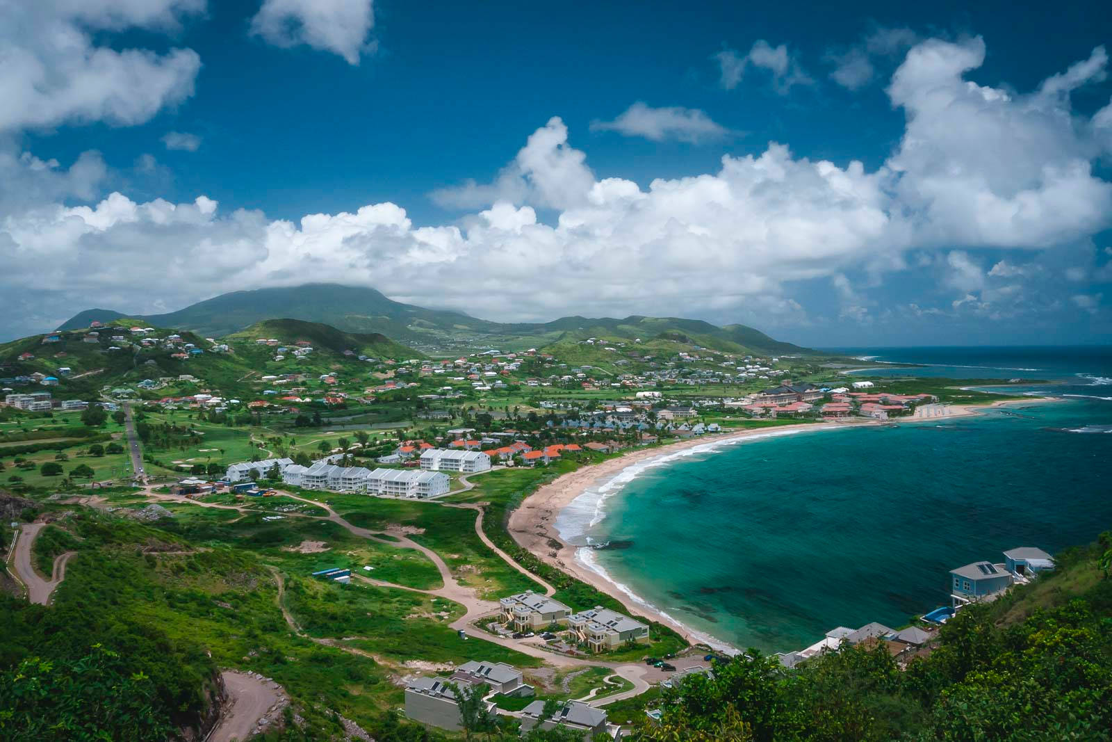 Caribbean 桌布 壁紙 — 一個充滿活力的沿海小鎮，坐落在鬱鬱蔥蔥的綠色山丘中，上方有碧綠的海水和白色的雲朵。