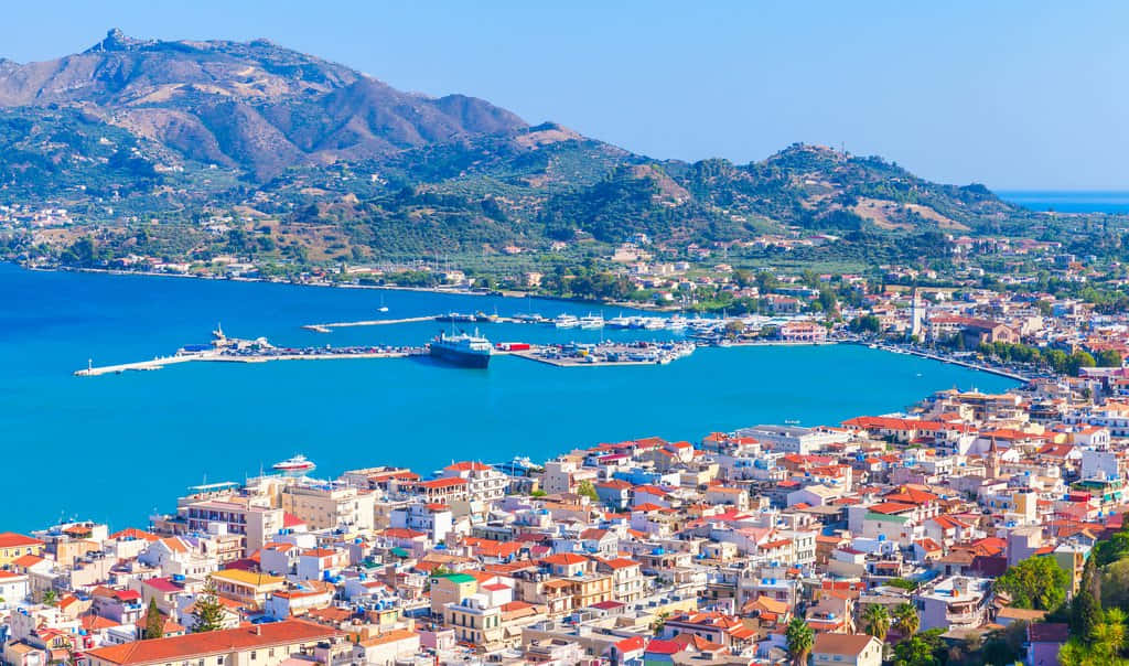 Greek Island 桌布 壁紙 — 一個充滿活力的沿海小鎮，擁有陶土色屋頂的建築，依偎在一個被陽光普照的山丘環繞的碧綠海灣旁，天空清澈湛藍。