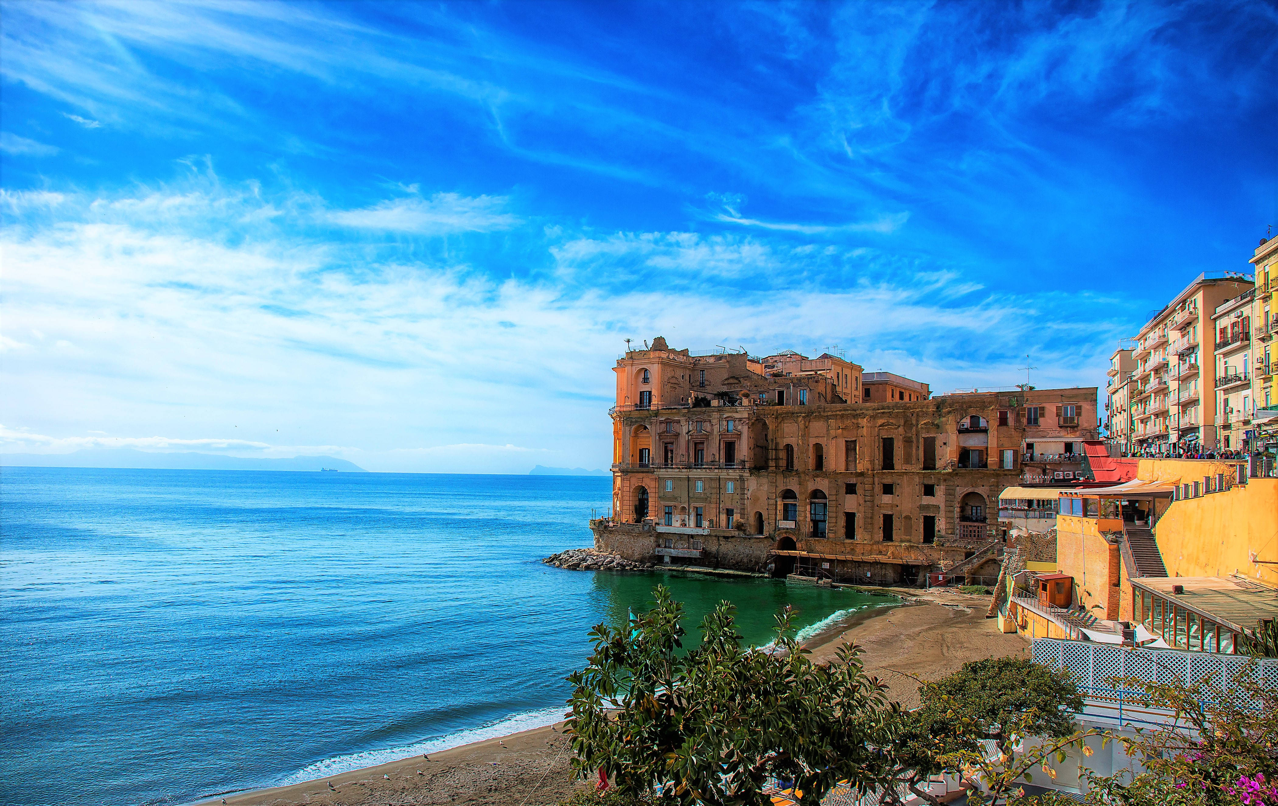 Naples 桌布 壁紙 — 一個充滿活力的沿海小鎮，歷史建築矗立在懸崖上，俯瞰著碧藍的海水，上方是明亮的藍天。