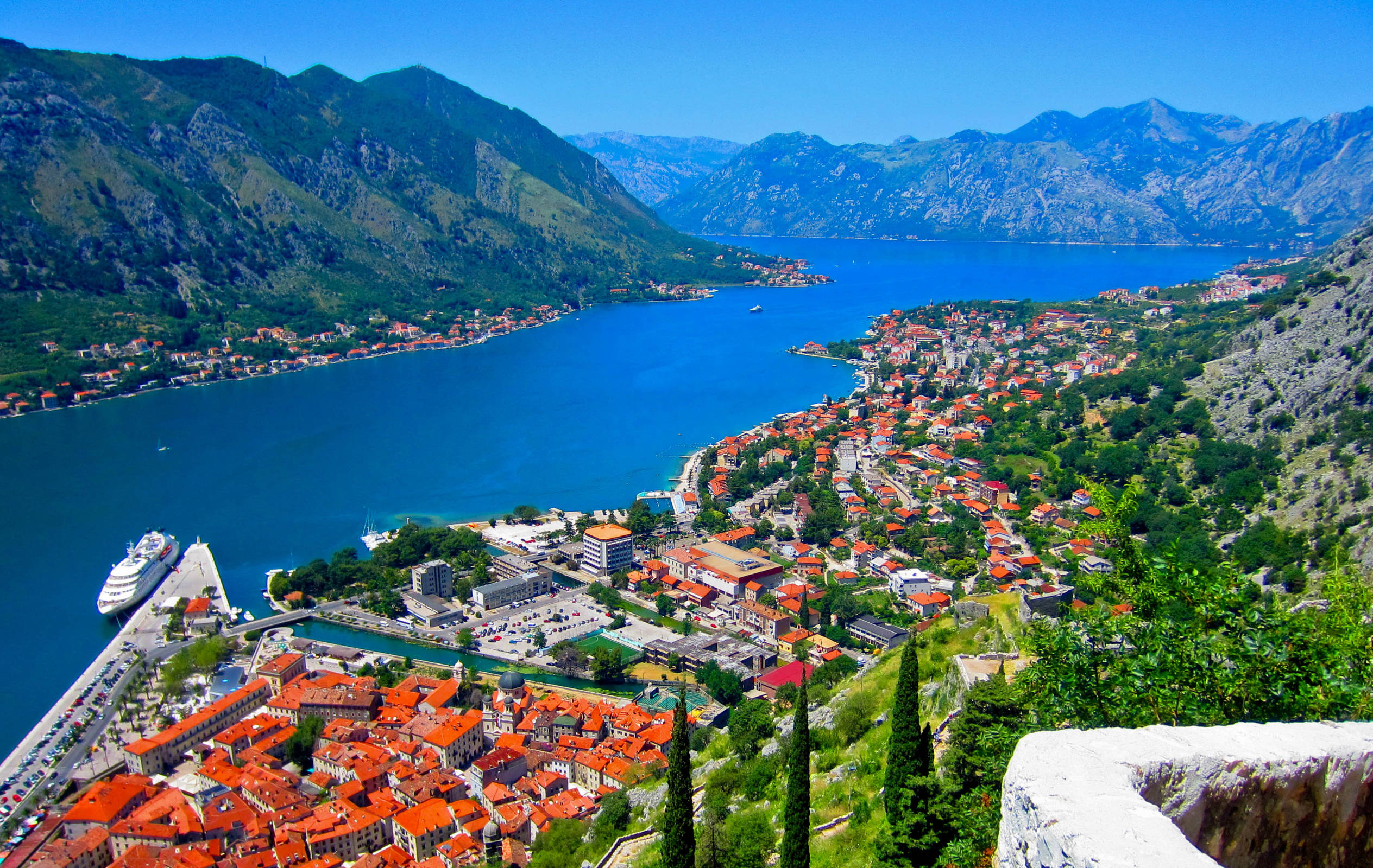 Montenegro 桌布 壁紙 — 一個充滿活力的沿海小鎮，陶土屋頂依附在綠色的山坡上，俯瞰著深藍色的海灣，四周環繞著高聳的山脈，天空晴朗。