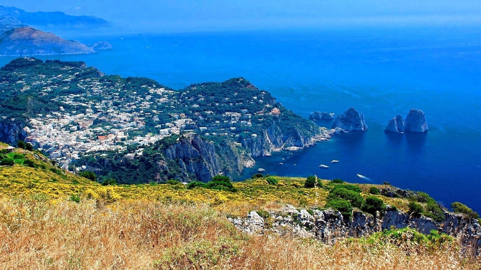 Capri 桌布 壁紙 — 一個充滿活力的沿海小鎮緊貼在陡峭的懸崖上，俯瞰深藍色的大海，遠處有標誌性的岩石 formations。