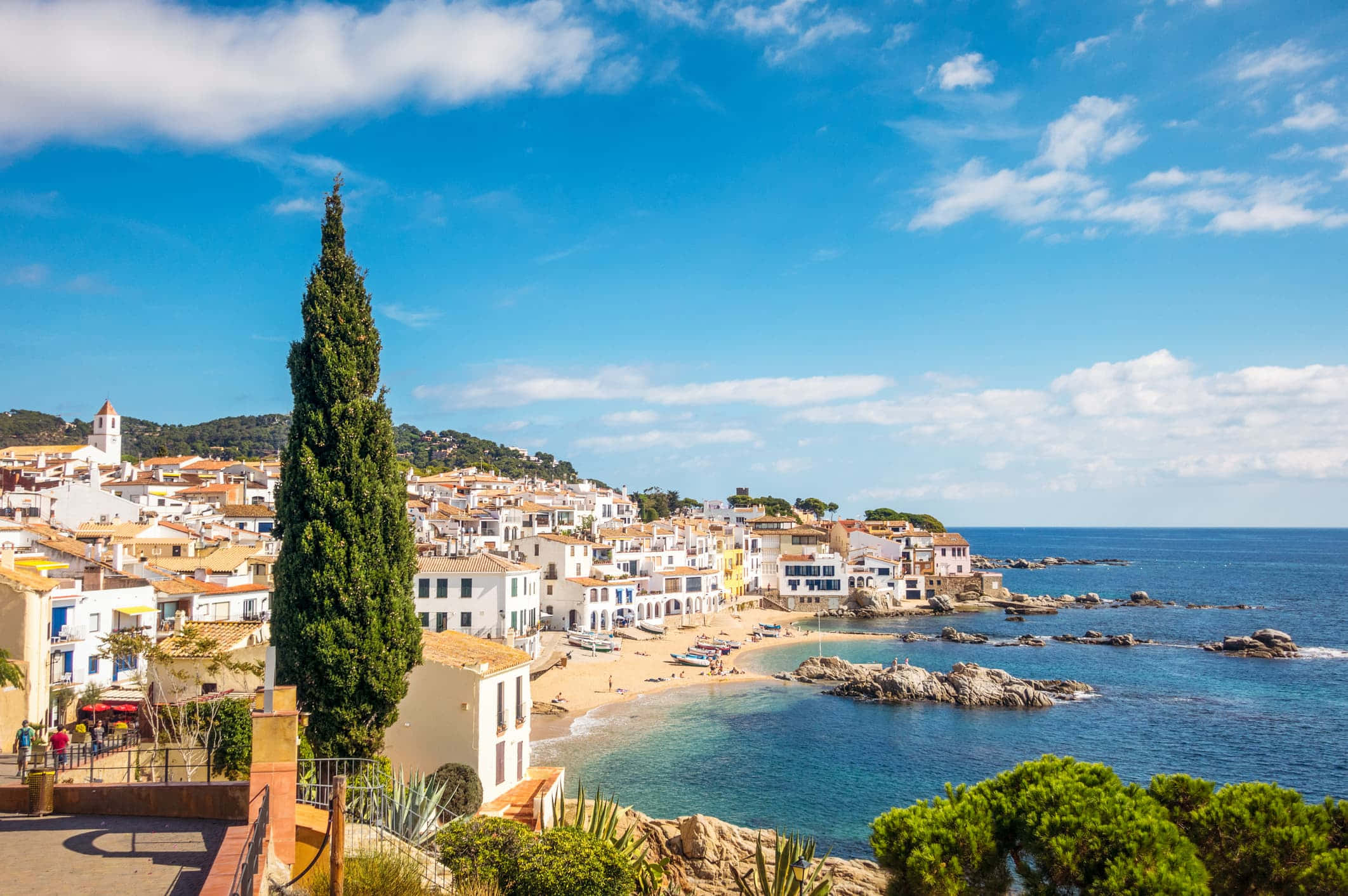 Spanish Beach 桌布 壁紙 — 一個充滿活力的沿海村莊，白色建築和陶土屋頂環抱著一個碧綠的海灣，上方是明亮的藍天，點綴著幾朵白雲。