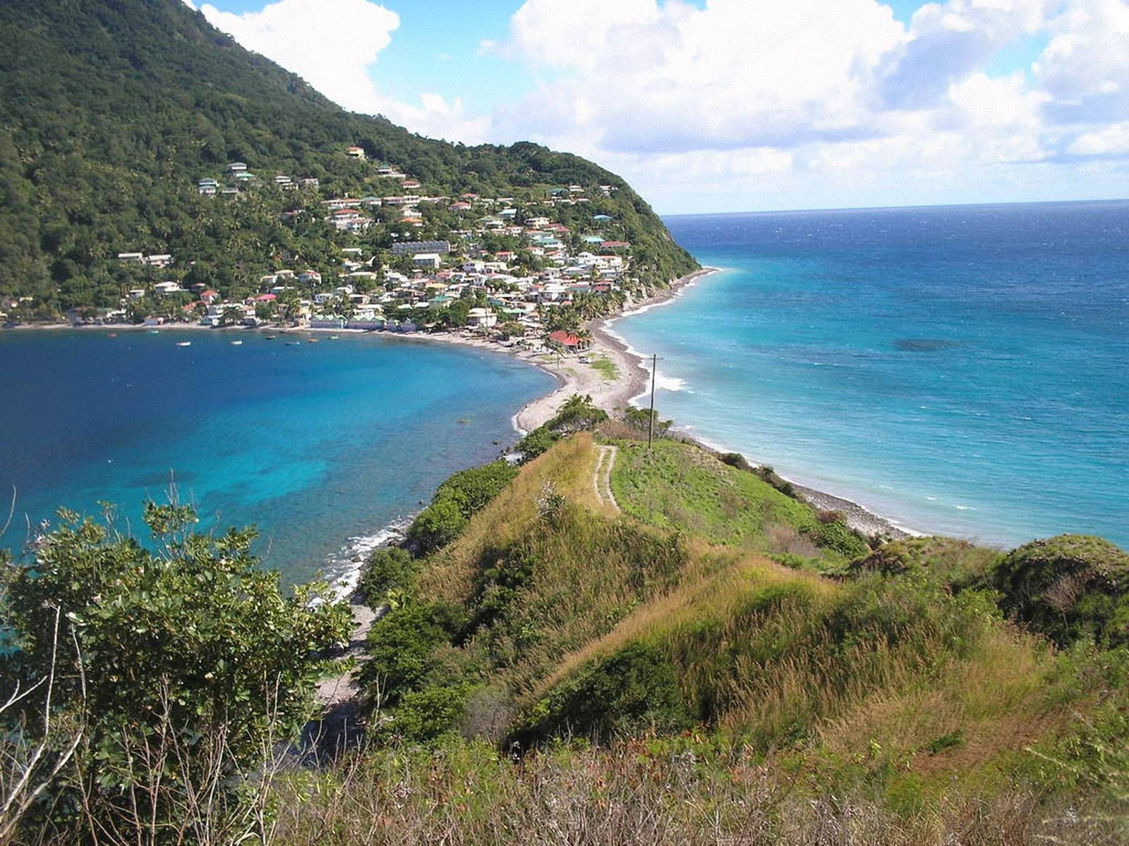 Dominica 桌布 壁紙 — 一個充滿活力的沿海村莊依附在鬱鬱蔥蔥的綠色山坡上，俯瞰著碧綠的海水，頭頂是明亮的藍天和蓬鬆的白雲。
