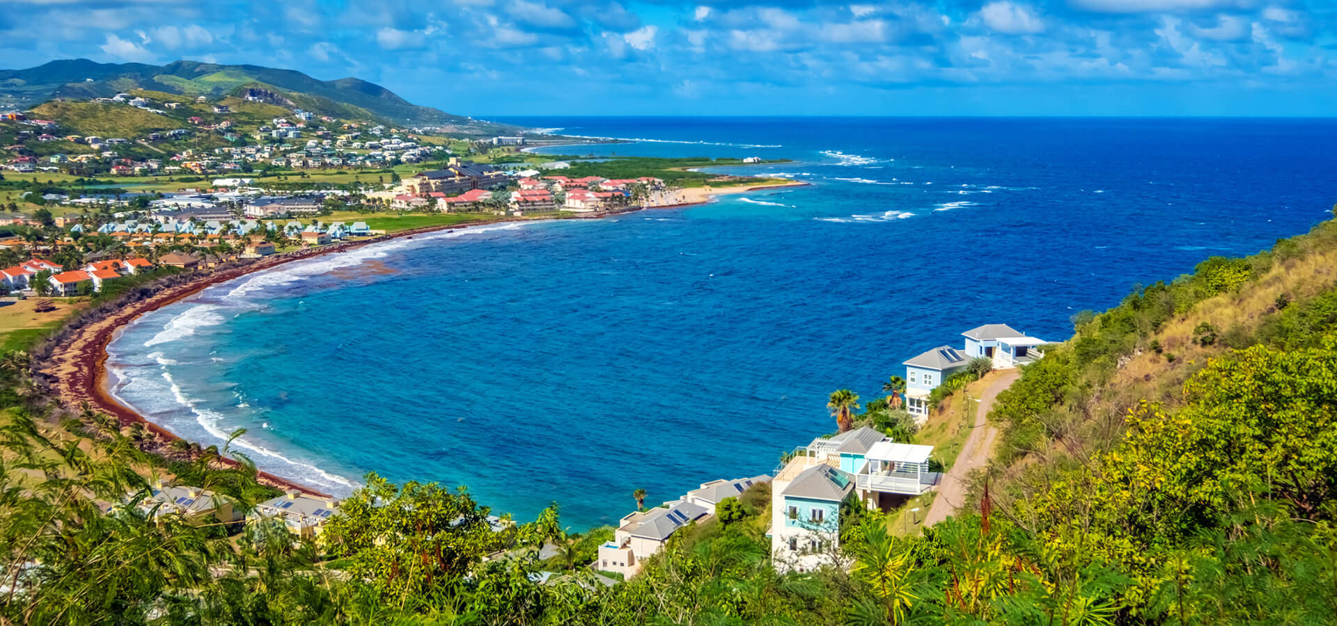 Caribbean 桌布 壁紙 — 一個充滿活力的海岸海灣，碧綠的海水環繞著沙灘，沙灘兩旁是色彩繽紛的房屋，坐落在鬱鬱蔥蔥的綠色山丘中，上方是明亮的藍天。
