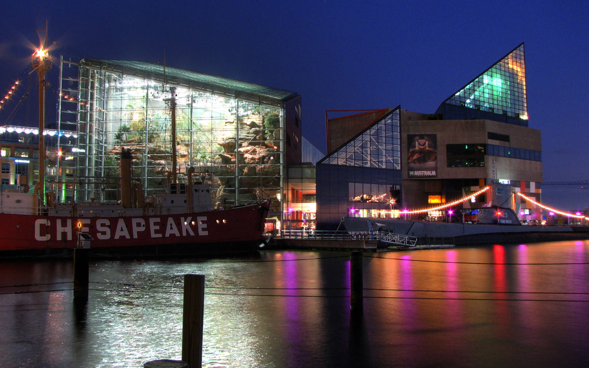 彩色動圖 桌布 壁紙 — 一個充滿活力的海濱夜景，一艘名為「CHESAPEAKE」的紅色船隻停泊在現代玻璃建築旁，建築物閃爍著五彩燈光，倒映在平靜的水面上。