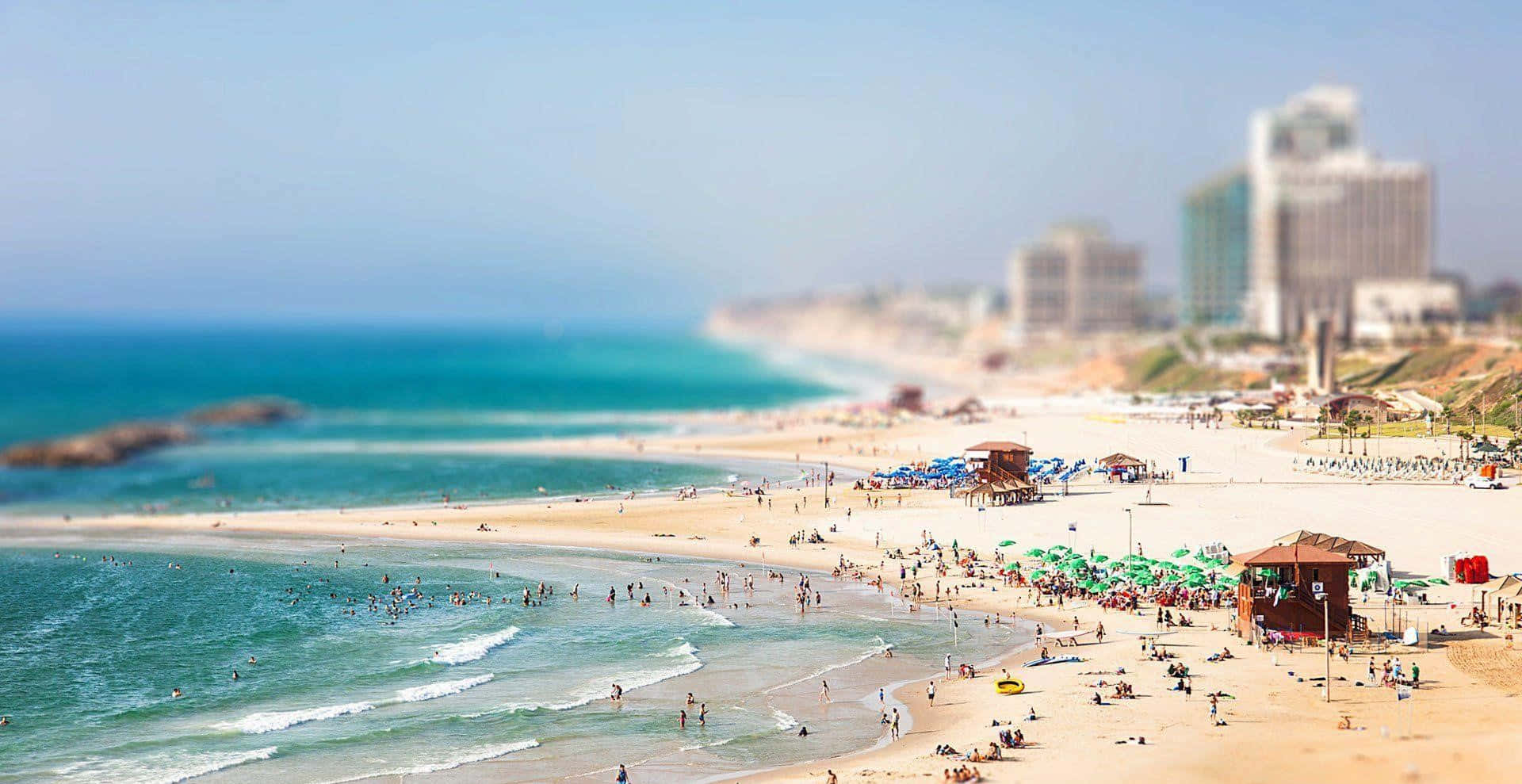 Israel 桌布 壁紙 — 一個充滿活力的海灘場景，青綠色的海浪拍打著擁擠的沙灘，沙灘上排列著色彩繽紛的遮陽傘和遠處的高樓大廈。