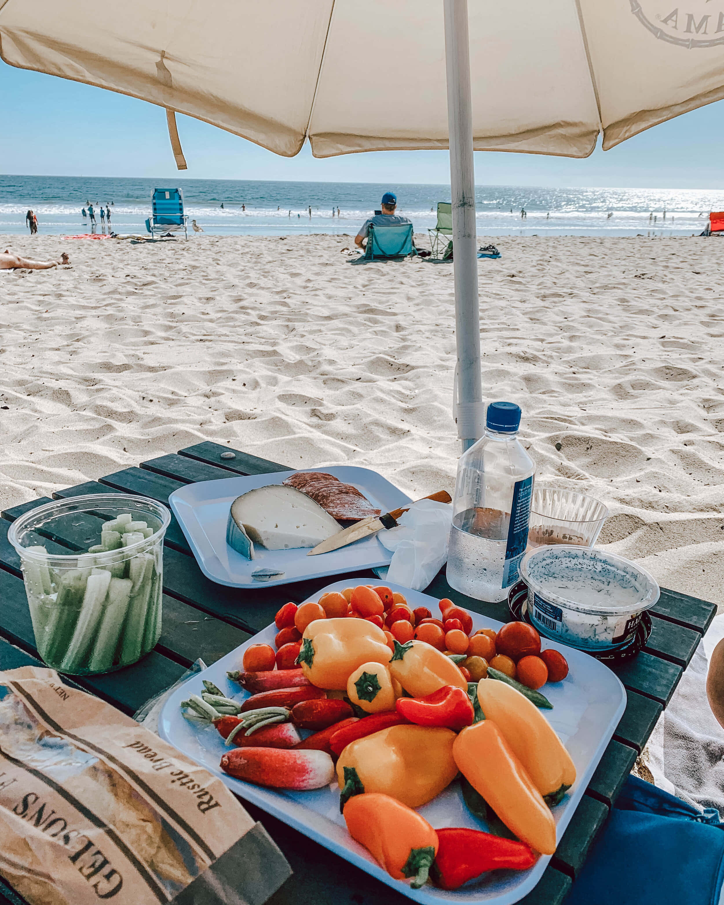 Beach Picnic 桌布 壁紙 — 一個充滿活力的海灘野餐，有色彩繽紛的甜椒、番茄和麵包，放在米色遮陽傘下，俯瞰海洋。