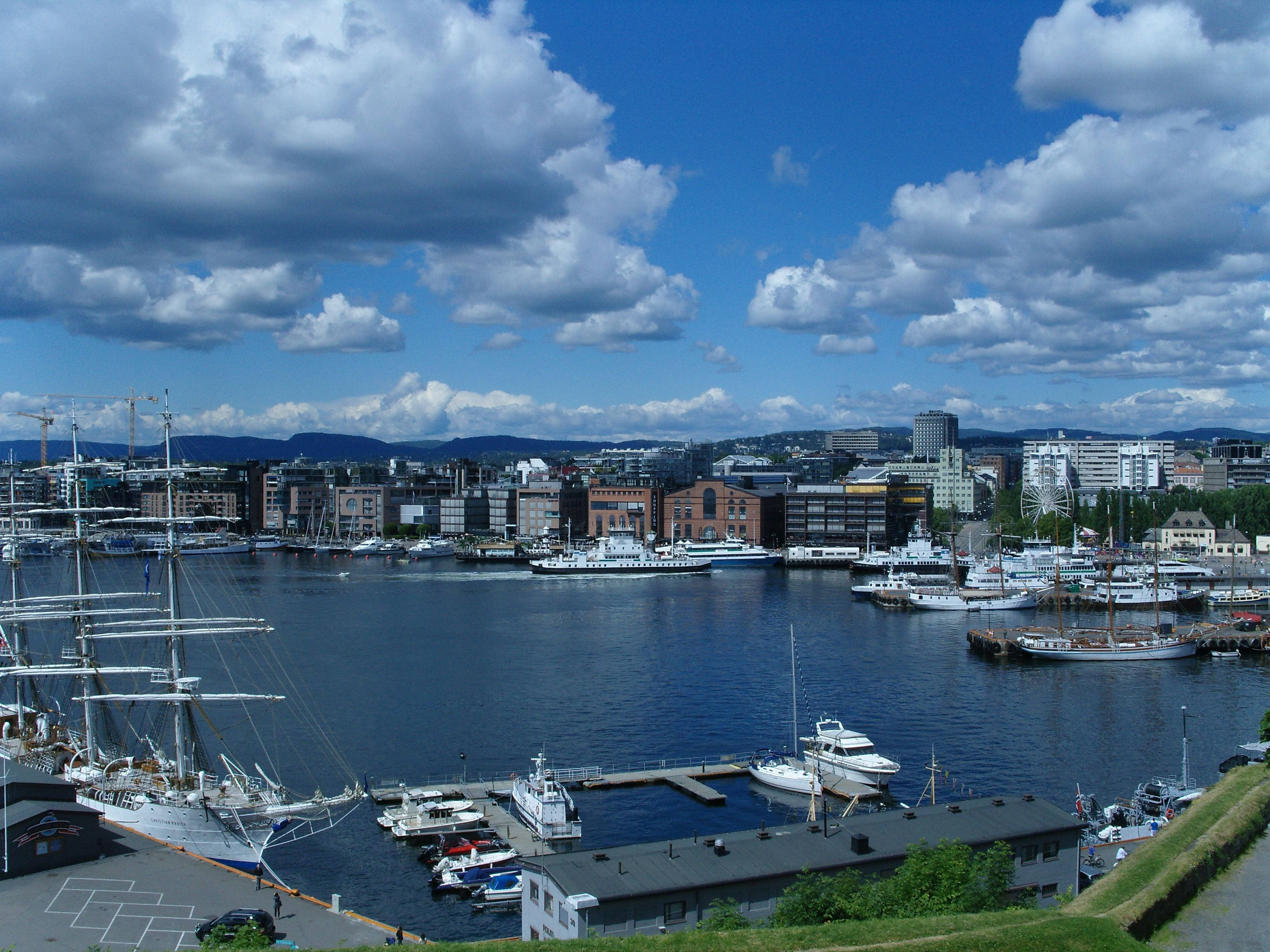 Norway 桌布 壁紙 — 一個充滿活力的港口城市景觀，有帆船和遊艇停泊在明亮的藍天下，伴隨著蓬鬆的白雲。