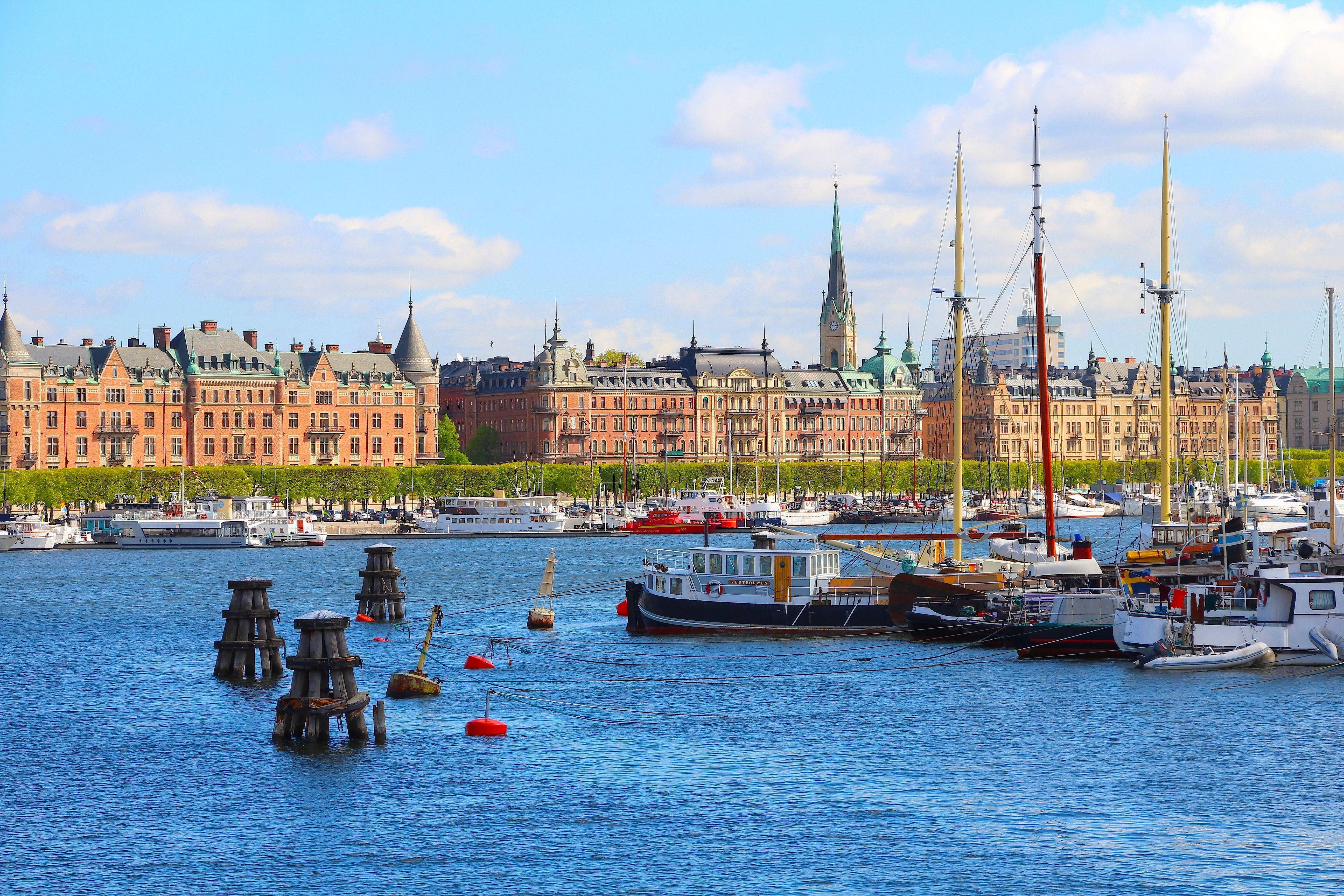 Sweden 桌布 壁紙 — 一個充滿活力的港口場景，擁有歷史悠久的磚砌建築、帆船和部分多雲天空下平靜的藍色海水。