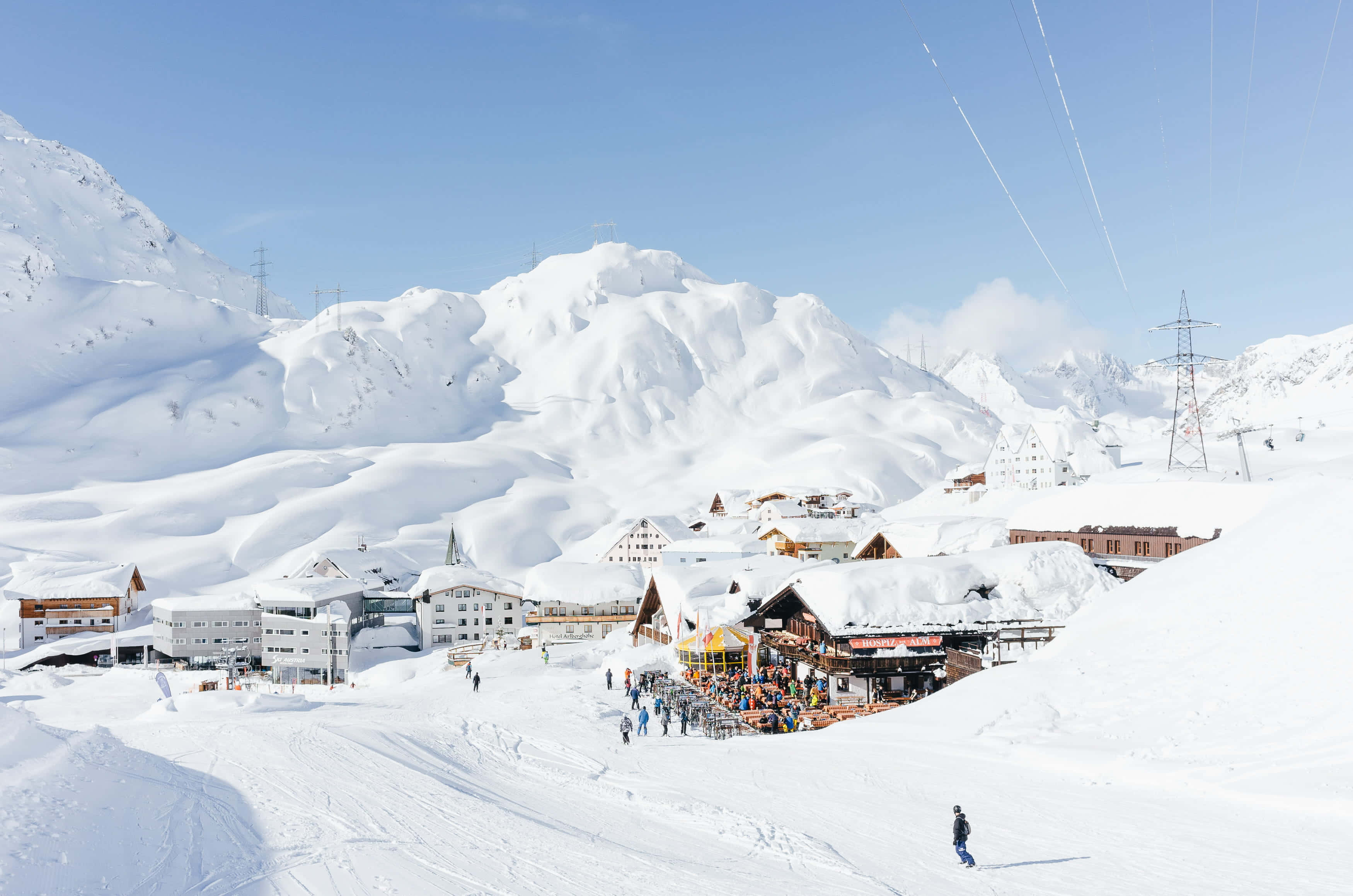 Ski Resort 桌布 壁紙 — 一個充滿活力的滑雪勝地，坐落在白雪覆蓋的山脈中，在晴朗的藍天下有滑雪者和木屋。