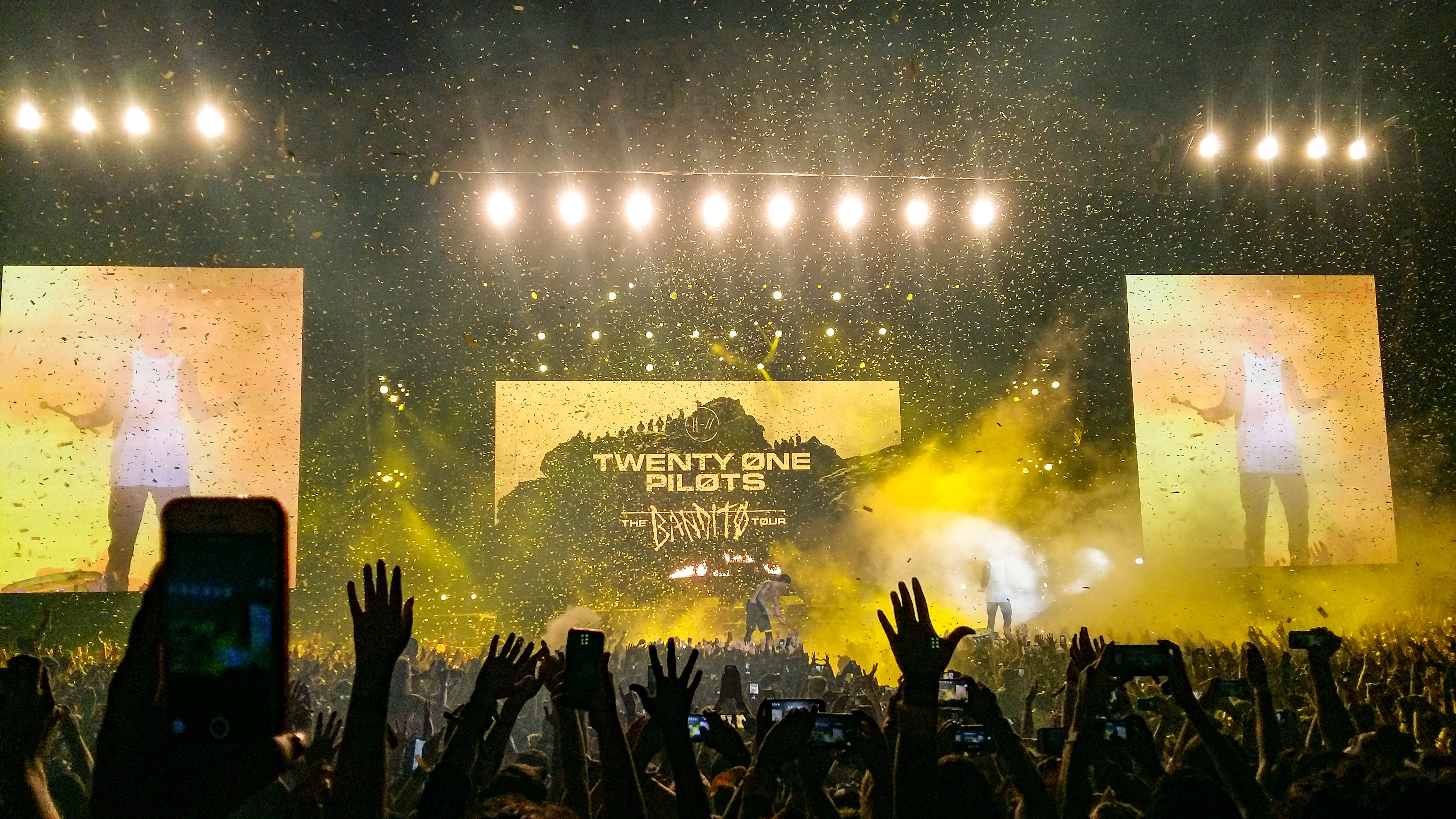 Lollapalooza 桌布 壁紙 — 一個充滿活力的演唱會場景，五彩紙屑飄落，粉絲們舉起手機和雙手，朝向燈光明亮的舞台上的Twenty One Pilots。