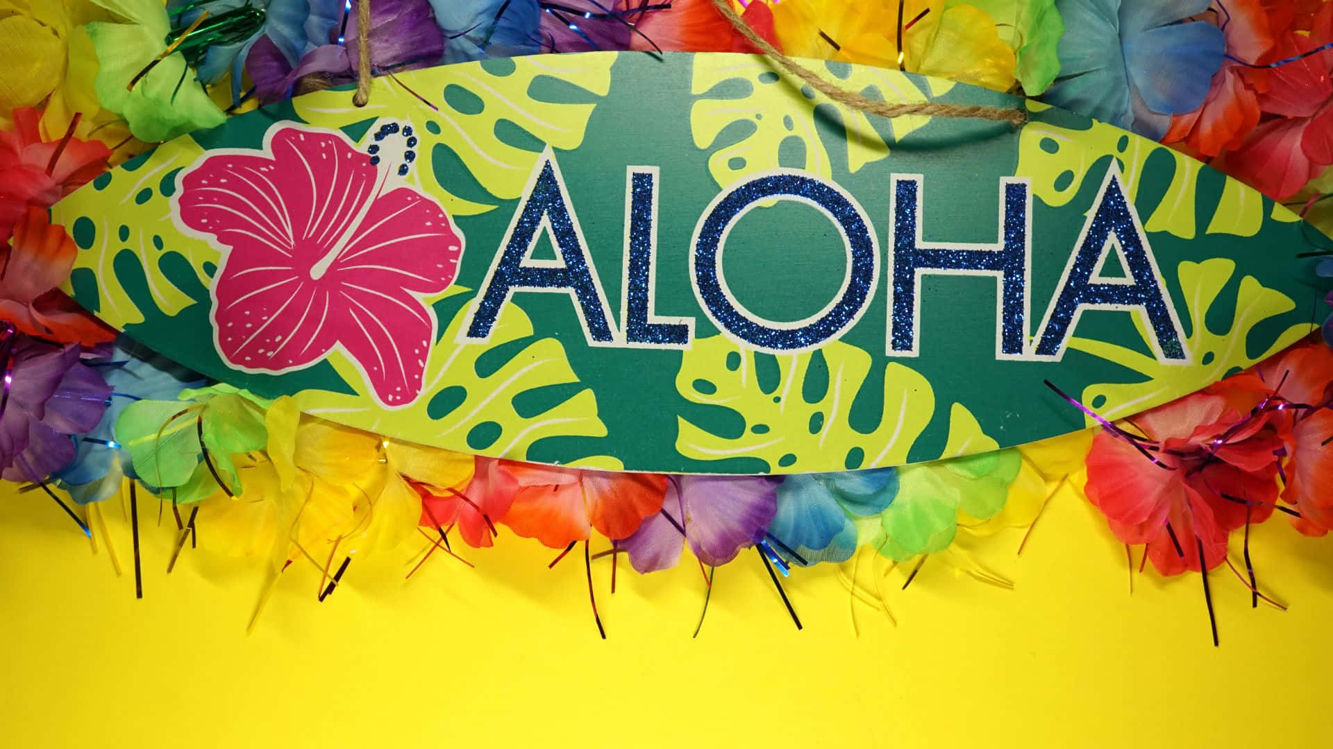 夏威夷 桌布 壁紙 — 一個充滿活力的熱帶標誌，帶有閃閃發光的「ALOHA」文字和粉紅色木槿，周圍環繞著色彩繽紛的羽毛花環，背景為明亮的黃色。