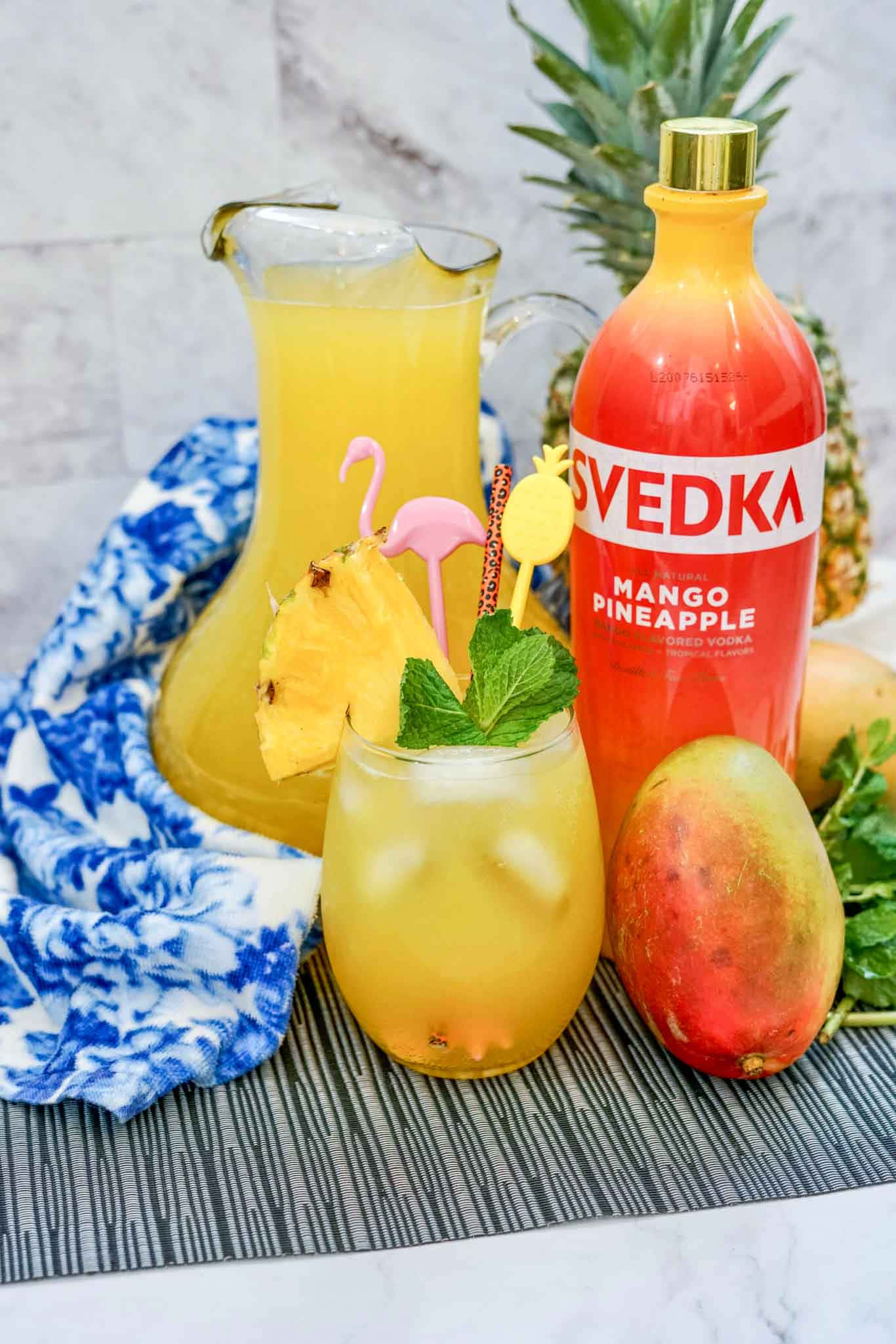 Mango 桌布 壁紙 — 一個充滿活力的熱帶飲品擺設，包括一瓶Svedka芒果鳳梨伏特加、一杯加冰和鳳梨裝飾的玻璃杯，以及圖案墊上的新鮮水果。