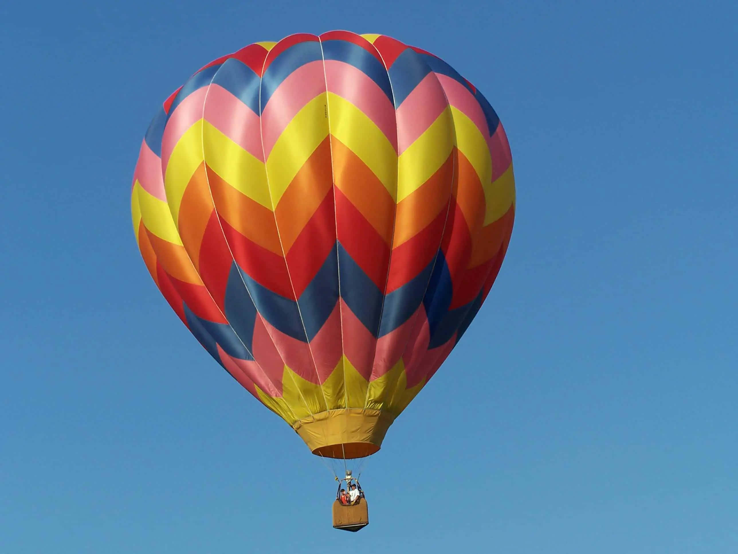 Hot Air Balloon 桌布 壁紙 — 一個充滿活力的熱氣球，帶有粉紅色、黃色、橙色和藍色的鋸齒條紋，在晴朗的藍天中漂浮。