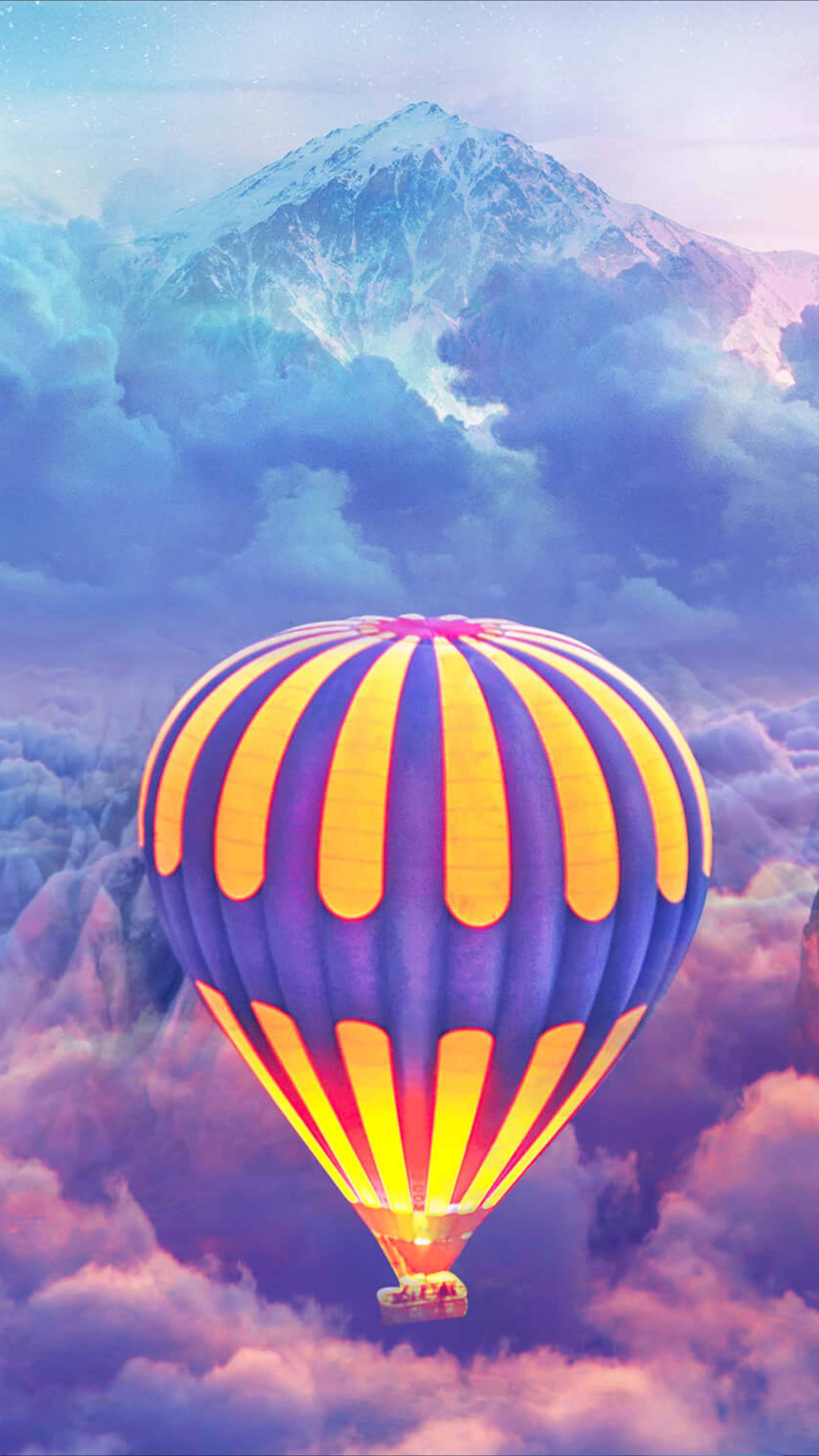 Hot Air Balloon 桌布 壁紙 — 一個充滿活力的熱氣球，帶有紫色和黃色條紋，漂浮在蓬鬆的雲層之上，朝著一座白雪覆蓋的山峰飛去，背景是柔和的天空。