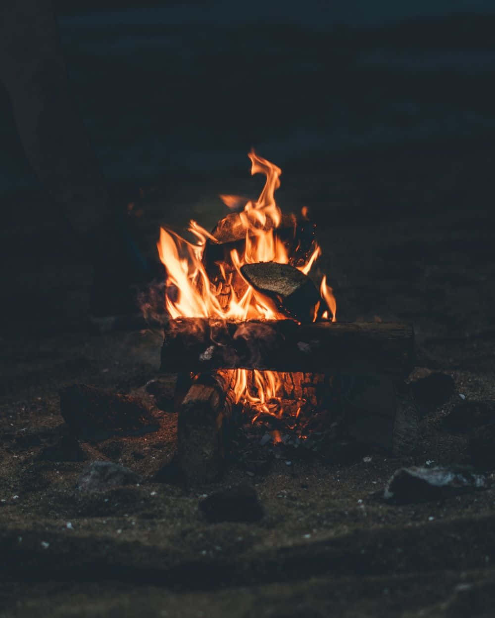 Beach Camping 桌布 壁紙 — 一個充滿活力的營火在漆黑的夜空背景下劈啪作響，橙色的火焰將溫暖的光芒投射在周圍的岩石和泥土上。