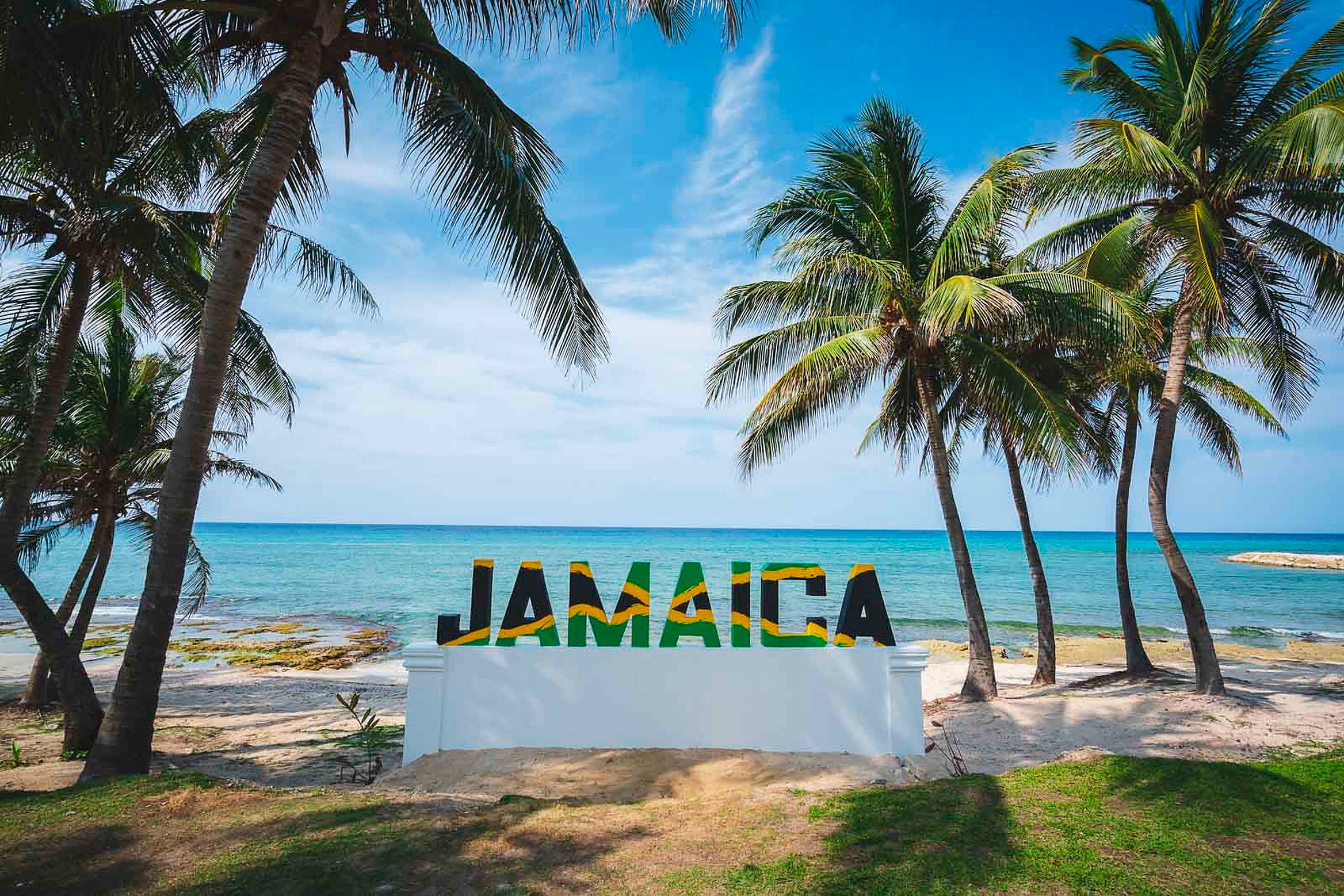 Jamaica Beach 桌布 壁紙 — 一個充滿活力的牙買加標誌以國家色彩矗立在陽光明媚的海灘上，周圍環繞著搖曳的棕櫚樹和碧綠的海洋。