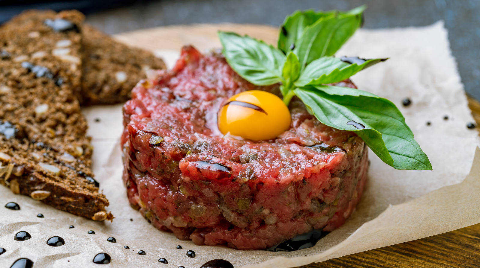 meat 桌布 壁紙 — 一個充滿活力的牛肉他他堆，上面放著一顆亮黃色的鵪鶉蛋和新鮮的羅勒葉，放在烘焙紙上。