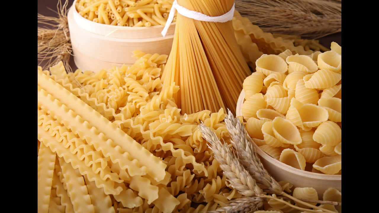 720p Pasta 桌布 壁紙 — 一個充滿活力的特寫，展示各種乾義大利麵類型，如義大利麵、螺旋麵和通心麵，與麥稈一起擺放在木碗中。