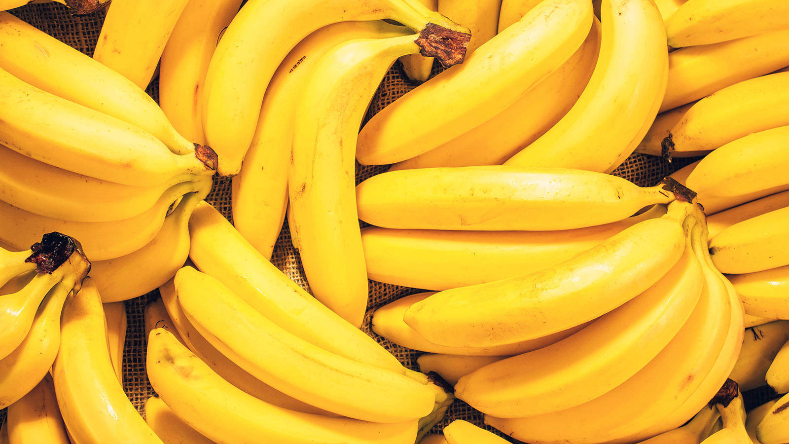 Plantain 桌布 壁紙 — 一個充滿活力的特寫，成熟的黃色香蕉堆積在帶有紋理的網狀表面上。