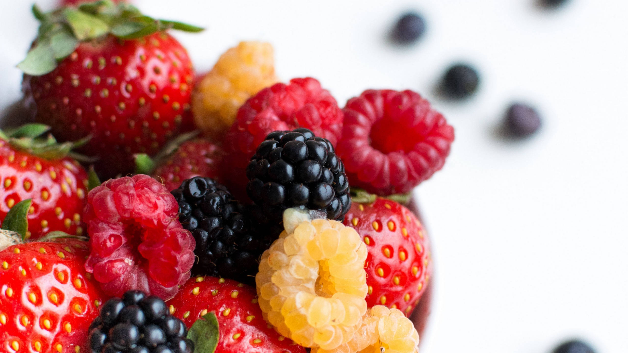 Berry 桌布 壁紙 — 一個充滿活力的特寫，新鮮草莓、覆盆子、黑莓和金黃覆盆子排列在白色表面上，背景散落著藍莓。