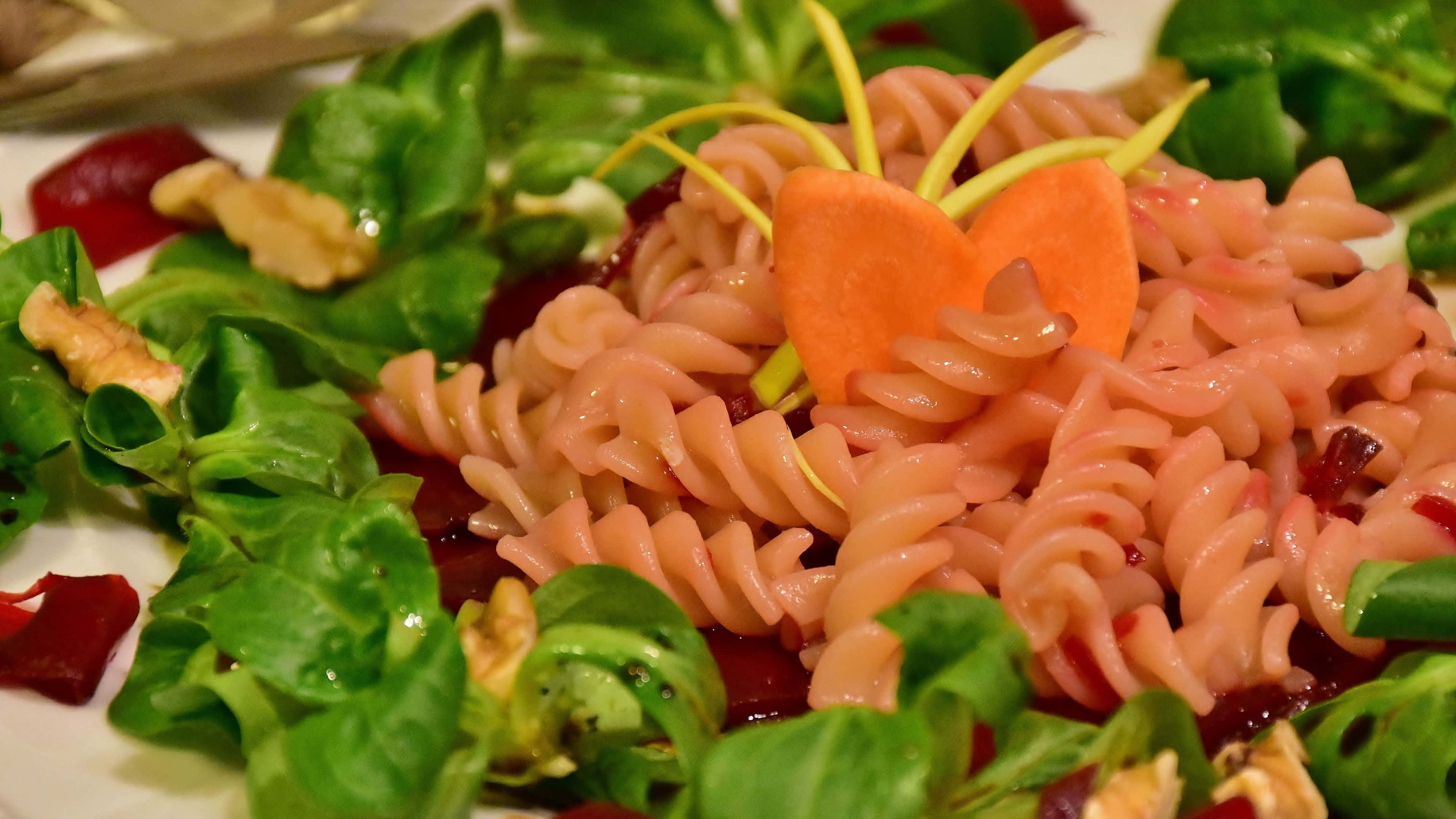 4K Pasta 桌布 壁紙 — 一個充滿活力的特寫，粉紅色螺旋義大利麵放在新鮮蔬菜中，配上胡蘿蔔裝飾和核桃碎。