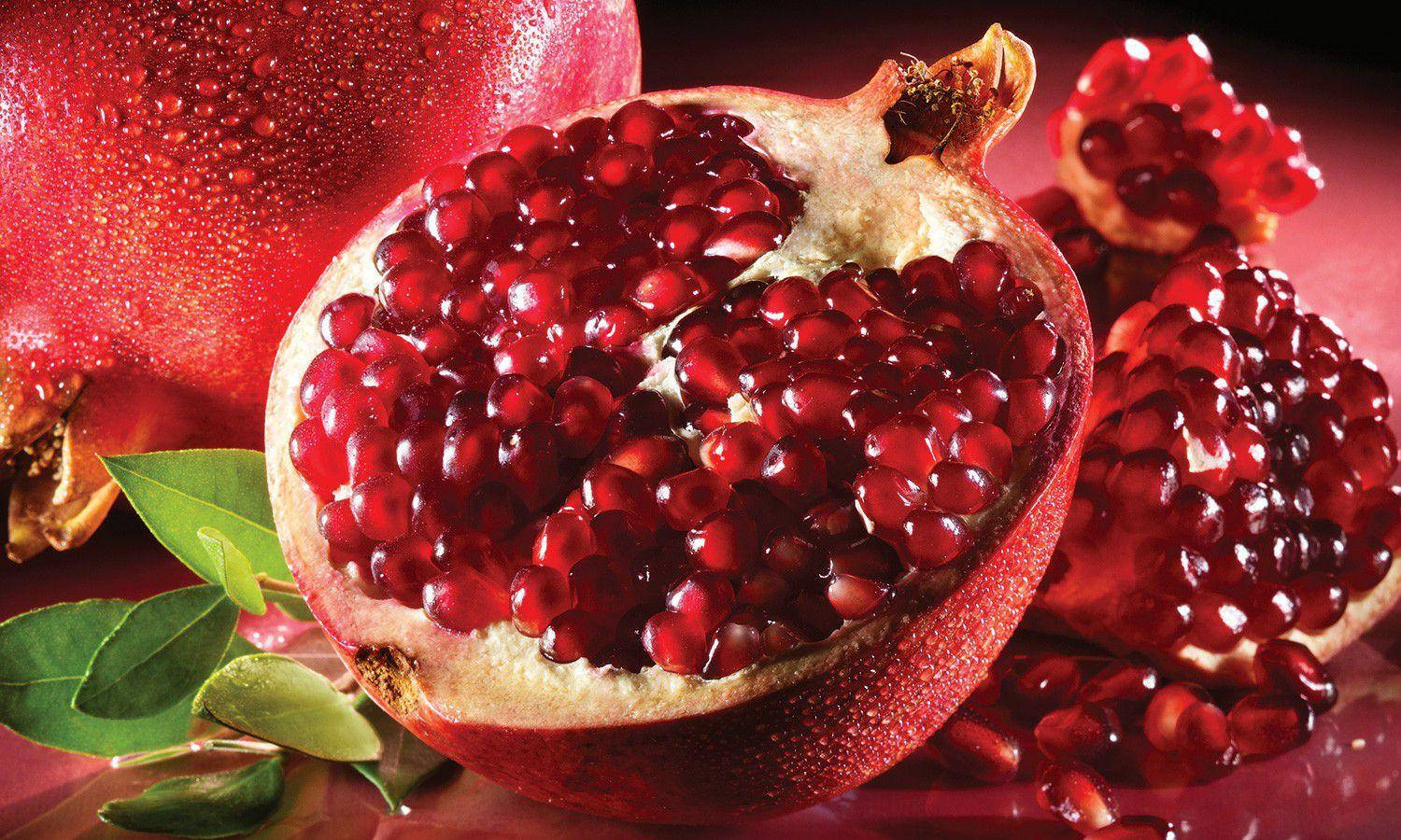 Fruits 桌布 壁紙 — 一個充滿活力的特寫鏡頭，展示了一個切半的石榴，露出多汁的紅寶石籽，旁邊是完整的水果和綠葉。