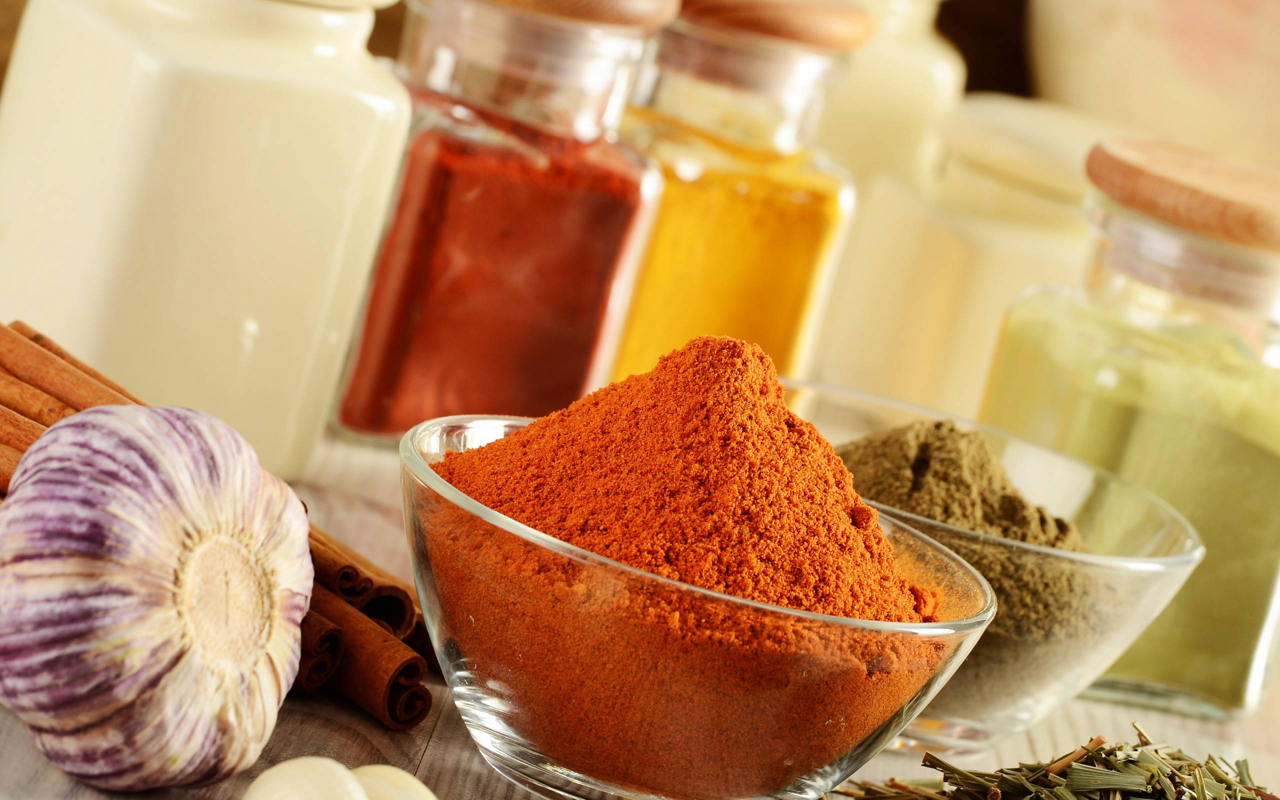 Spice 桌布 壁紙 — 一個充滿活力的特寫鏡頭，展示包括紅粉、肉桂、大蒜和香草在內的香料，排列在木質表面上的玻璃罐和碗中。