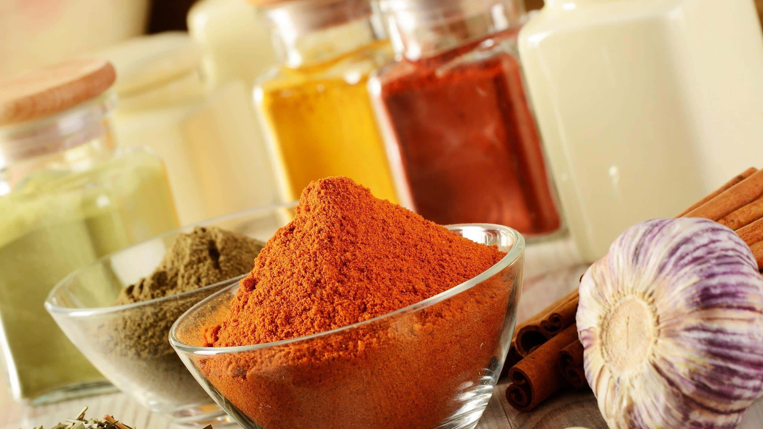 Spices 桌布 壁紙 — 一個充滿活力的特寫鏡頭，玻璃碗中裝滿了鮮紅色的辣椒粉，旁邊是整顆大蒜和肉桂棒。