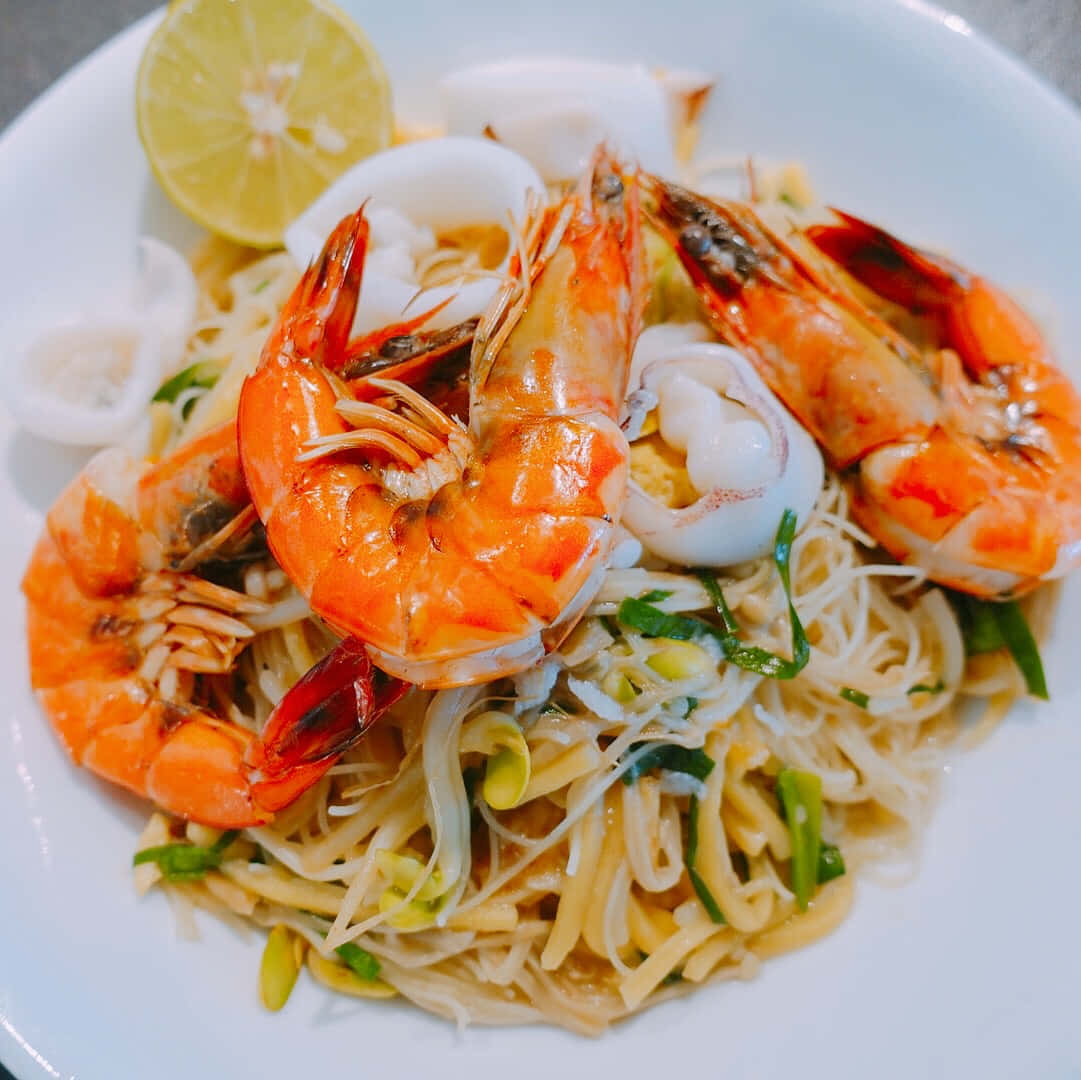 Noodles 桌布 壁紙 — 一個充滿活力的特寫鏡頭，白色盤子上堆滿了冒煙的麵條，上面鋪著鮮橙色的蝦和嫩魷魚圈，配以新鮮香草和萊姆角裝飾。