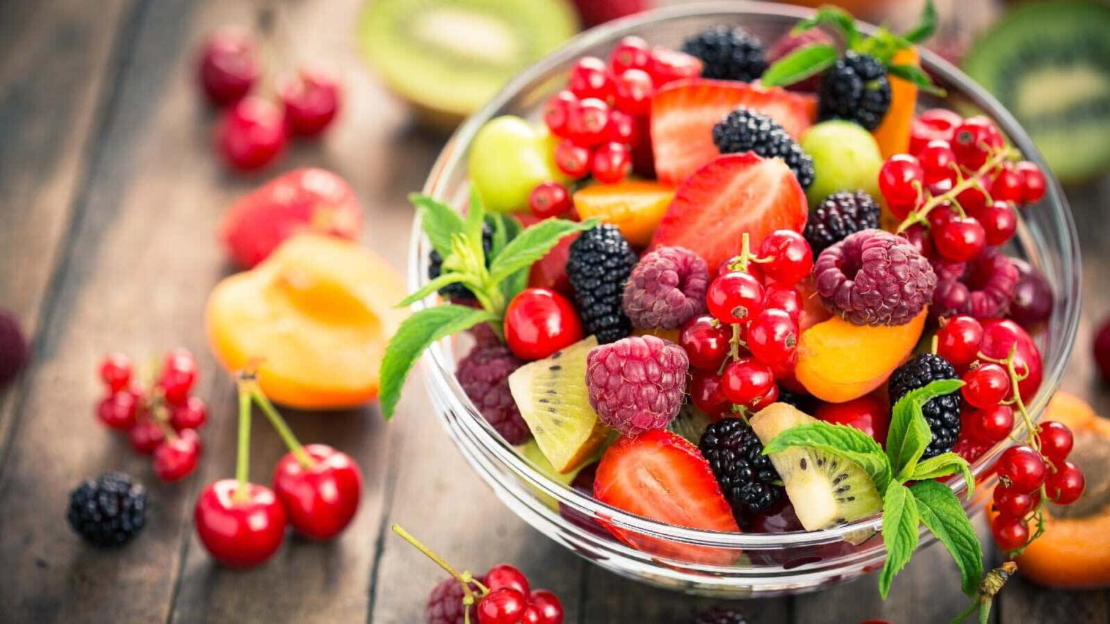 Fruit 桌布 壁紙 — 一個充滿活力的玻璃碗，裝滿新鮮漿果、櫻桃、奇異果和薄荷，放在質樸的木桌上。