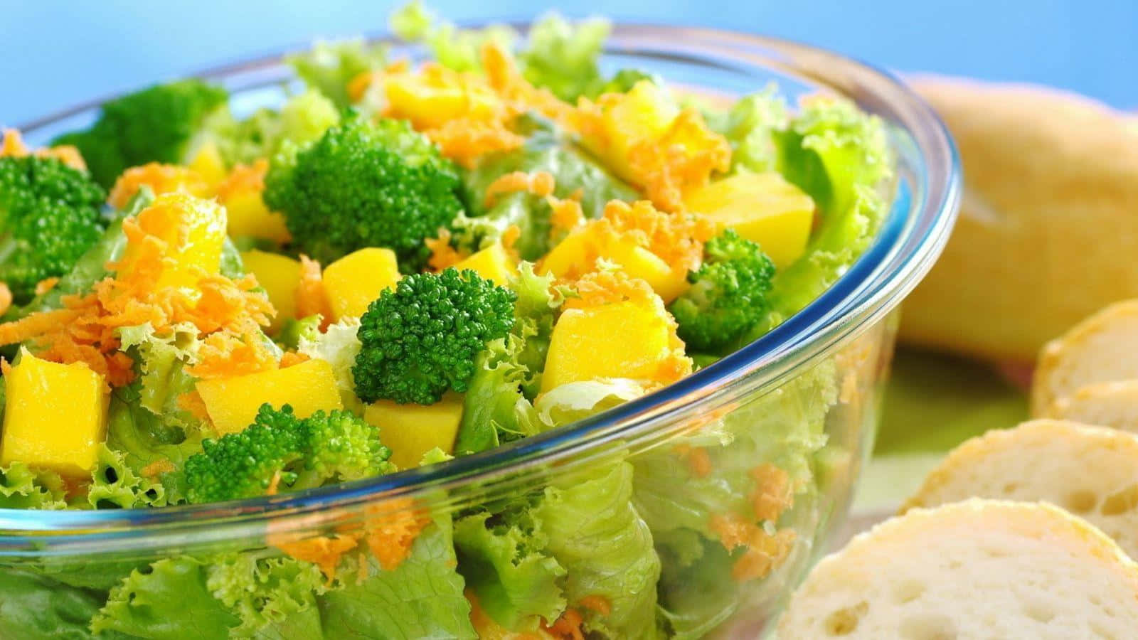 Healthy Food 桌布 壁紙 — 一個充滿活力的玻璃碗，裝滿新鮮的綠色生菜、西蘭花塊、芒果和刨絲紅蘿蔔，旁邊有切片麵包。