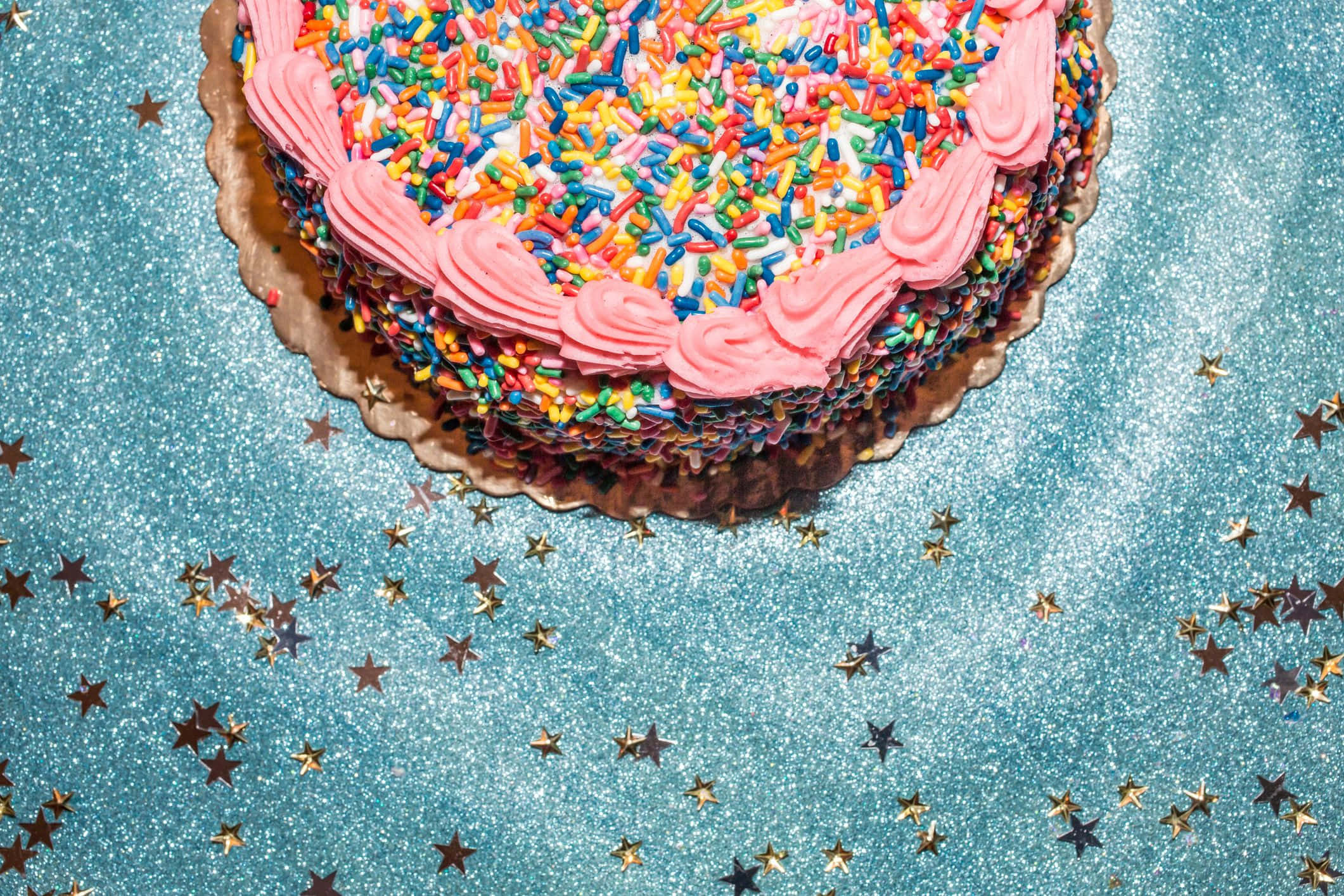 Birthday Cake 桌布 壁紙 — 一個充滿活力的生日蛋糕，帶有粉紅色糖霜和彩虹糖針，放置在一個散落著金色星星的閃亮藍色表面上。
