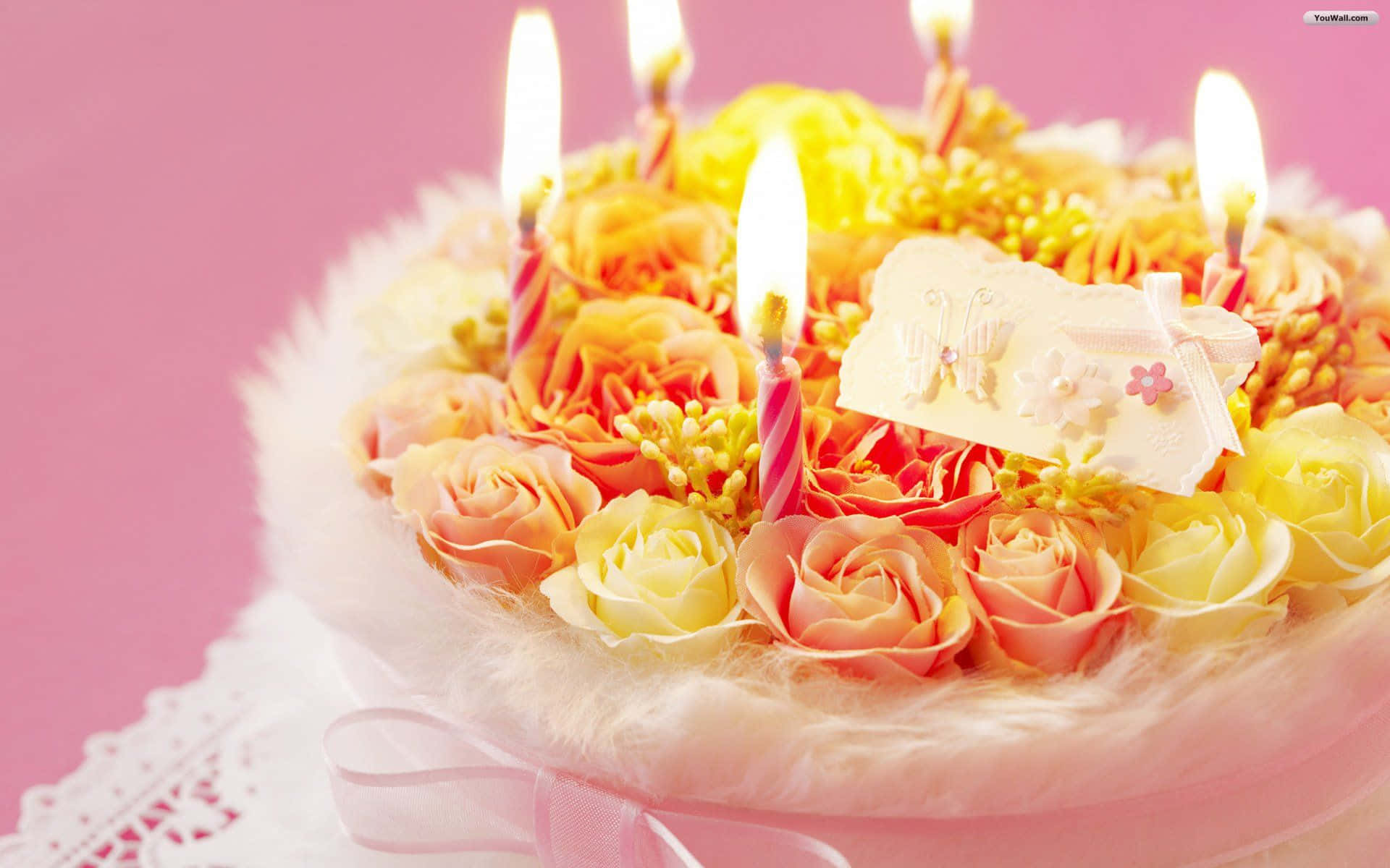 Birthday 桌布 壁紙 — 一個充滿活力的生日蛋糕，飾有蜜桃黃玫瑰、點燃的蠟燭和一個裝飾標籤，背景是柔和的粉紅色。