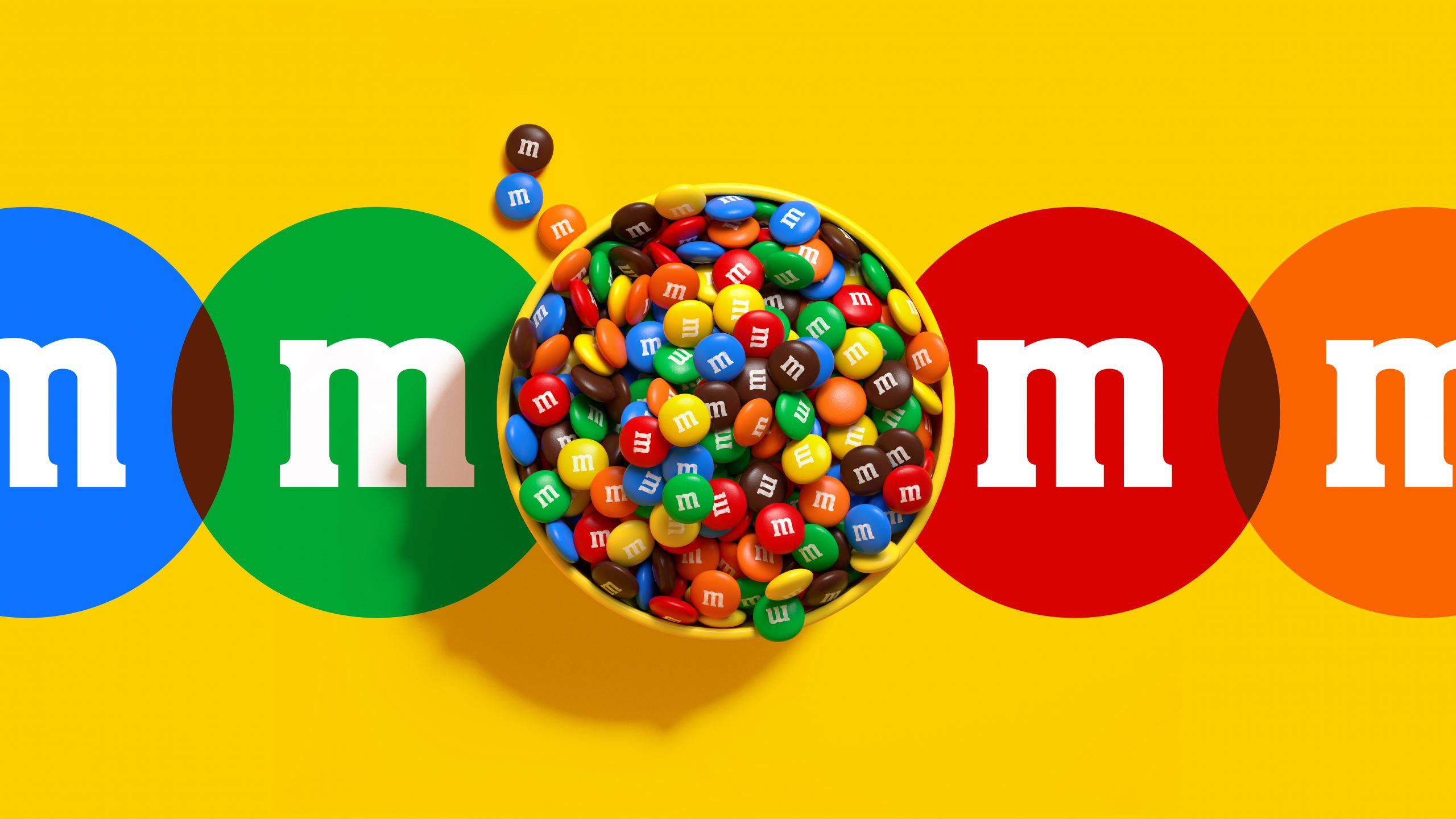 巧克力 桌布 壁紙 — 一個充滿活力的碗，裝滿了色彩繽紛的M&M's糖果，放在明亮的黃色背景上，兩側有大型品牌圓圈。