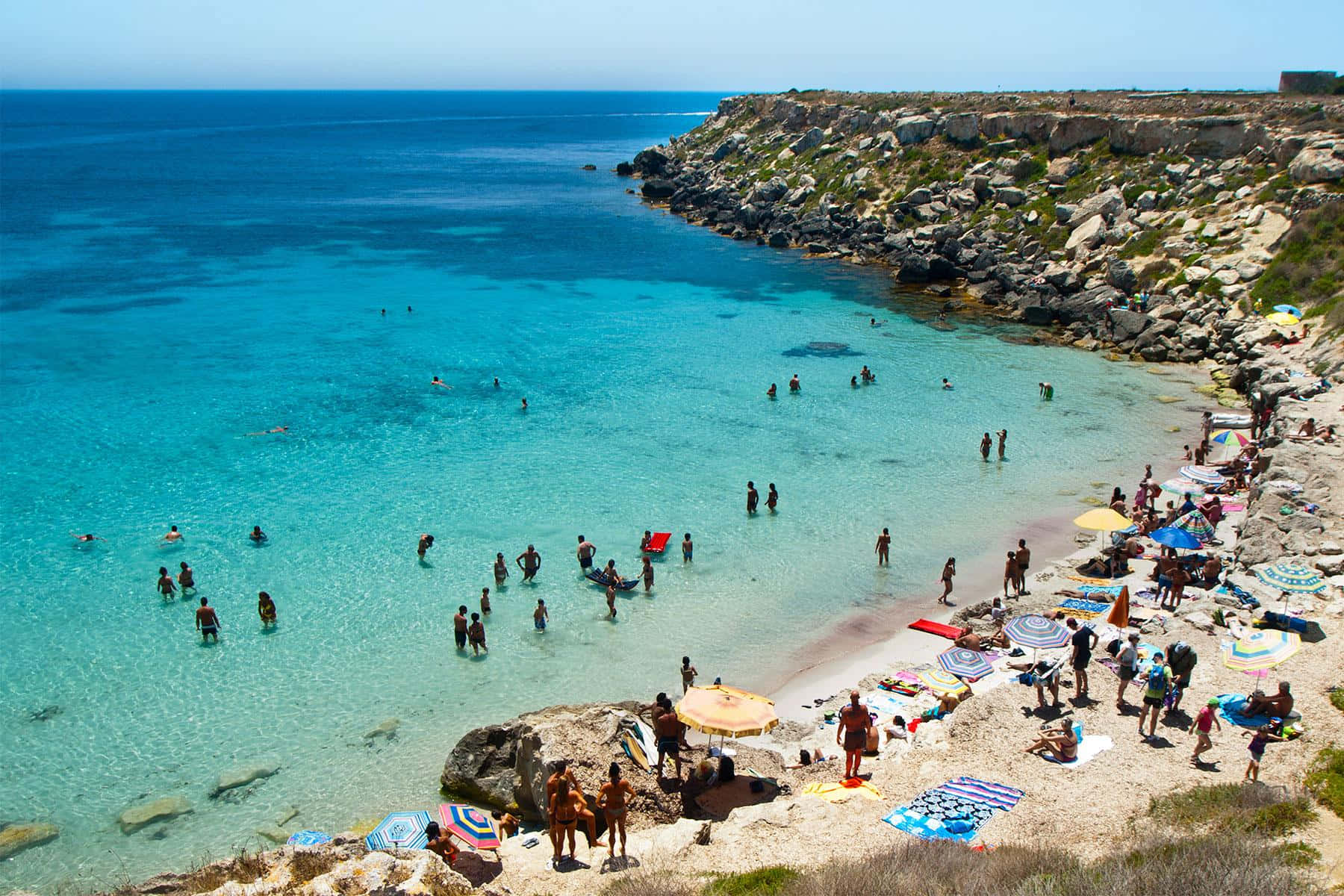 Italian Beach 桌布 壁紙 — 一個充滿活力的碧綠海灣，在清澈的藍天下擠滿了享受日光浴和游泳的人們。