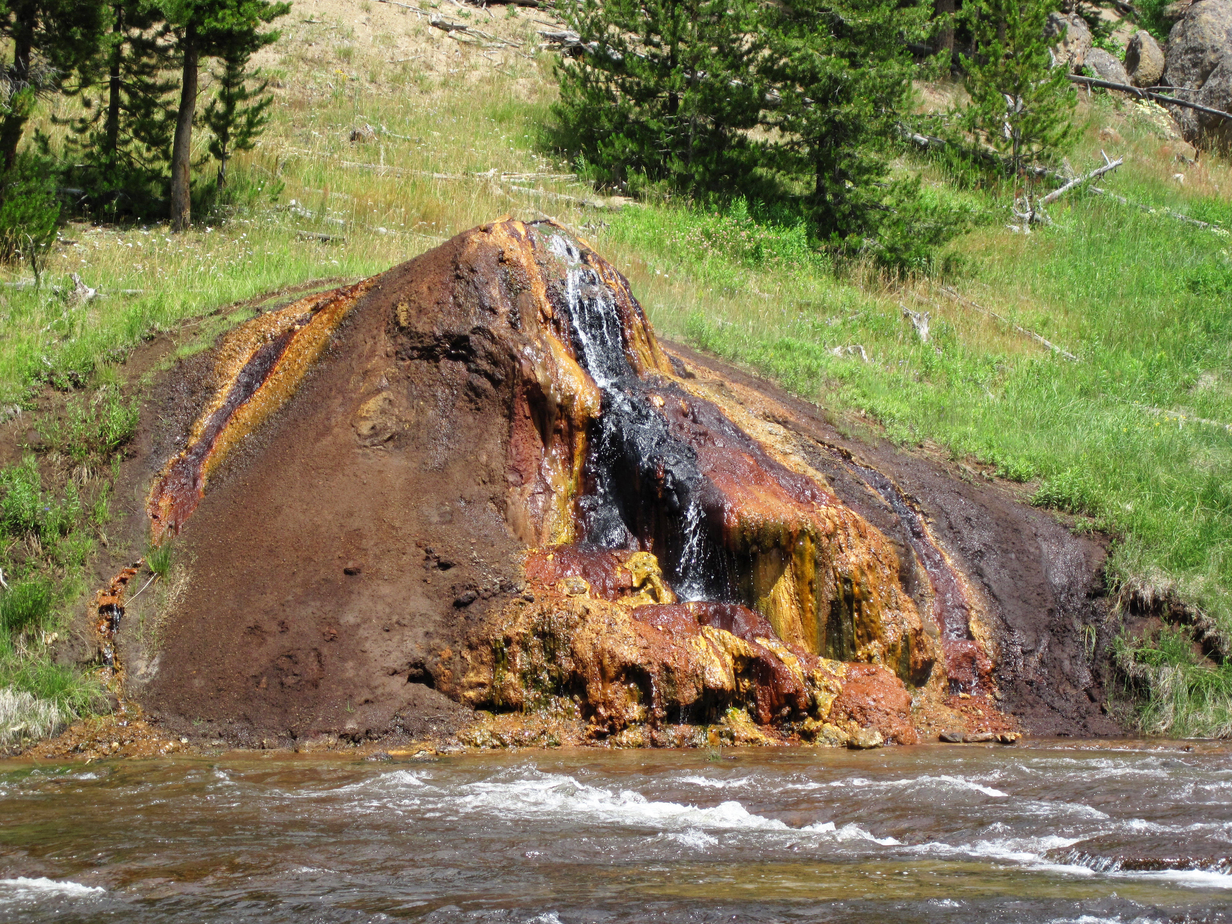 Yellowstone 桌布 壁紙 — 一個充滿活力的礦物溫泉沿著生鏽的岩石層層疊疊地流入一條湍急的河流，周圍環繞著綠草和松樹。