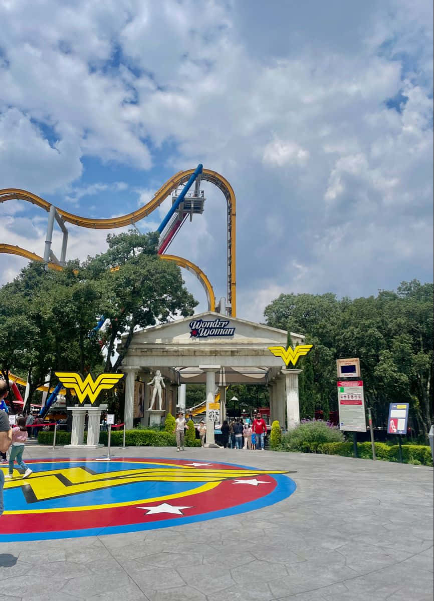 Six Flags 桌布 壁紙 — 一個充滿活力的神力女超人主題雲霄飛車聳立在宏偉的入口上方，客人在部分多雲的天空下，走近地上色彩繽紛的標誌。