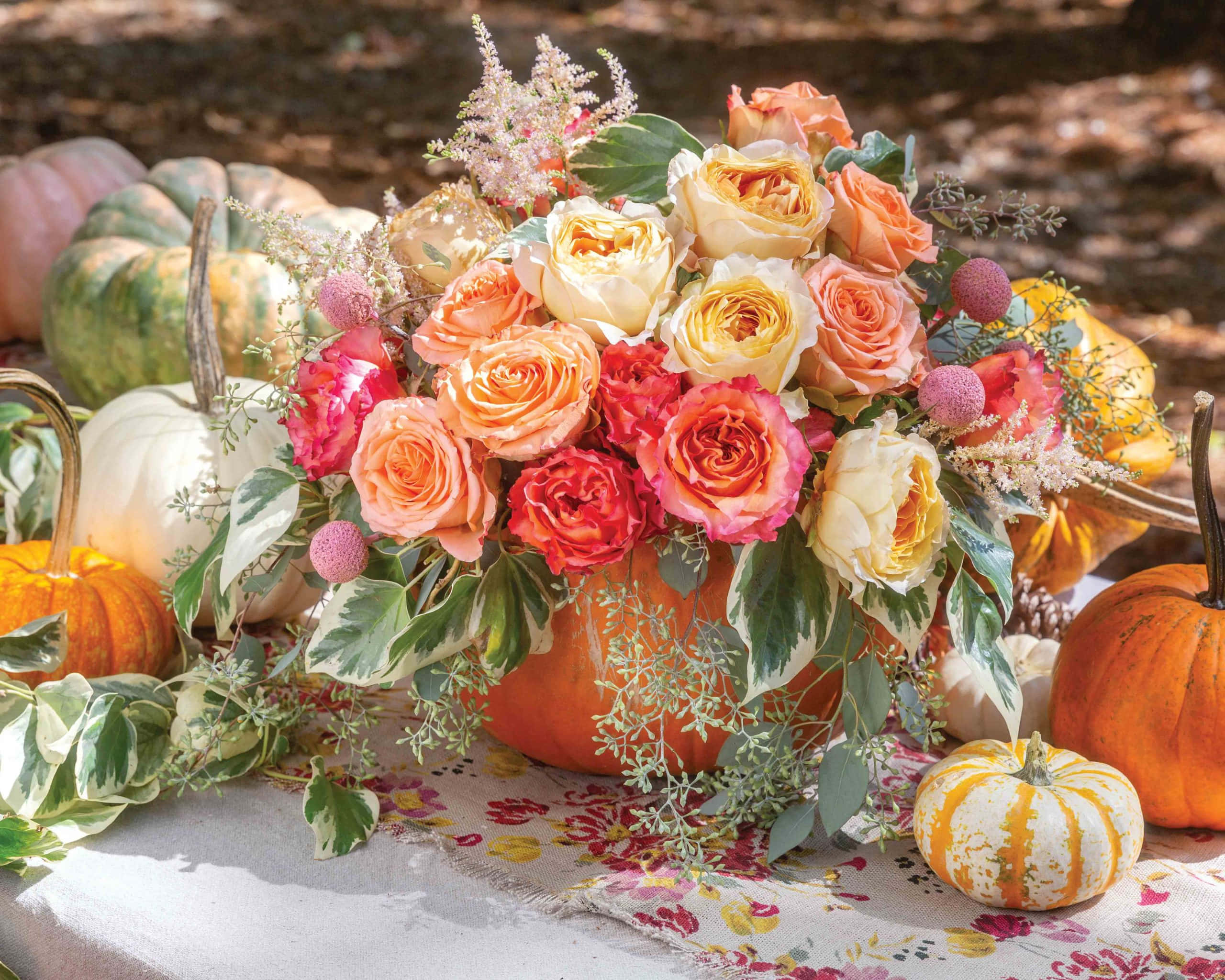Autumn Bounty 桌布 壁紙 — 一個充滿活力的秋季花卉佈置，放在南瓜花瓶裡，周圍有各式各樣的南瓜和樹葉，放在有圖案的桌布上。