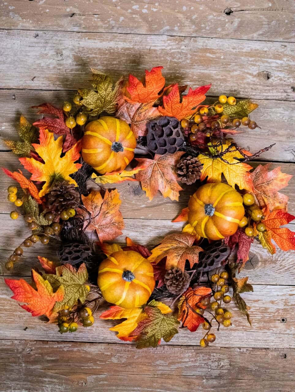 Fall Wreath 桌布 壁紙 — 一個充滿活力的秋季花環，裝飾著橙色南瓜、楓葉、松果和漿果，放置在風化的木板上。