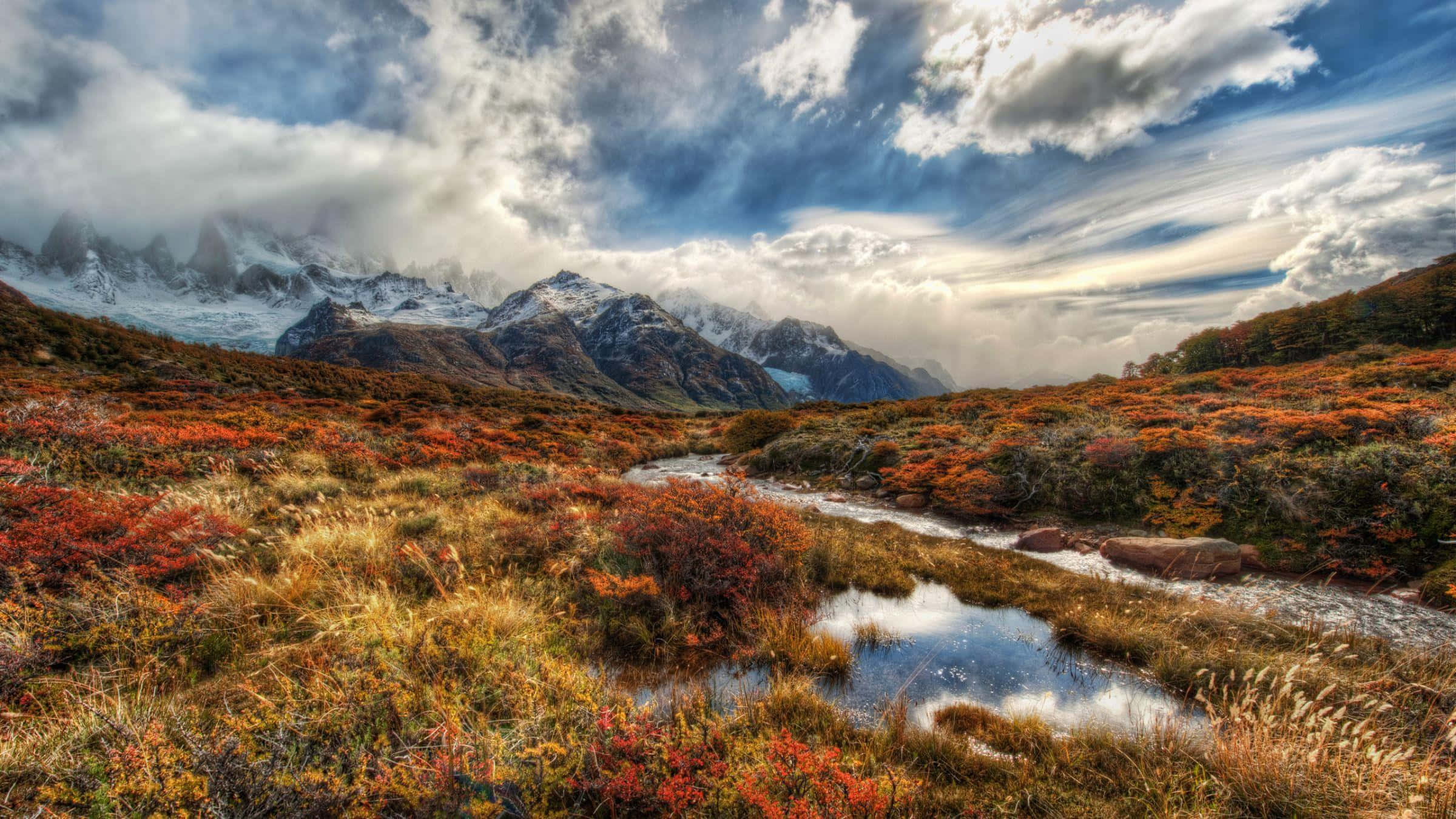 Patagonia 桌布 壁紙 — 一個充滿活力的秋日山谷，蜿蜒的小溪映照著雪山上空戲劇性的天空。