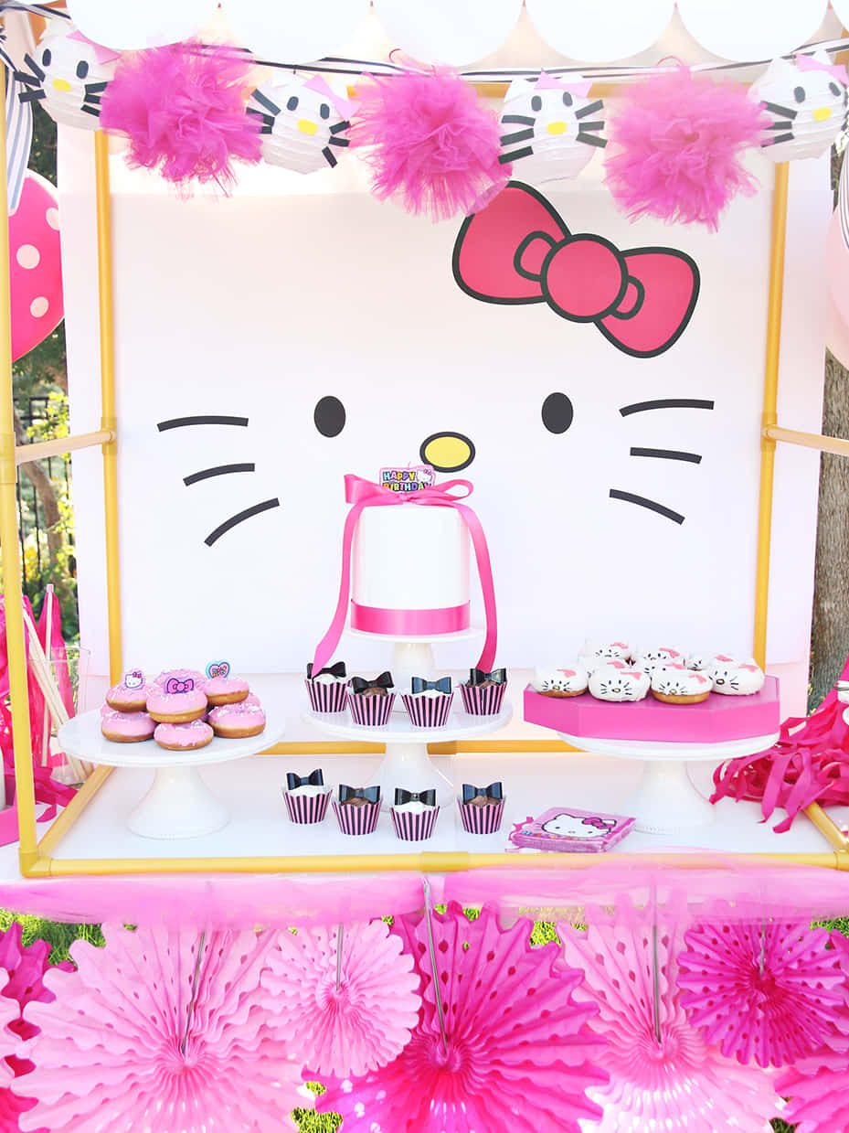 Hello Kitty Party 桌布 壁紙 — 一個充滿活力的粉紅色Hello Kitty甜點桌，上面有杯子蛋糕、甜甜圈和一個蛋糕，背景是一個巨大的卡通面孔。