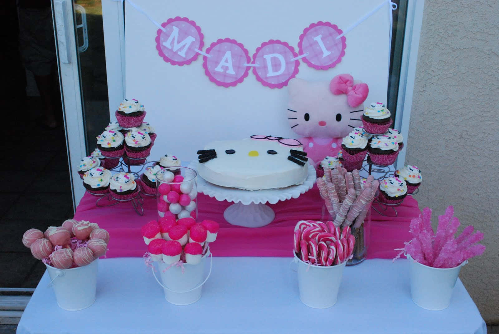 Hello Kitty Birthday 桌布 壁紙 — 一個充滿活力的粉紅色Hello Kitty生日甜點桌，在名字橫幅下擺放著杯子蛋糕、蛋糕和糖果。